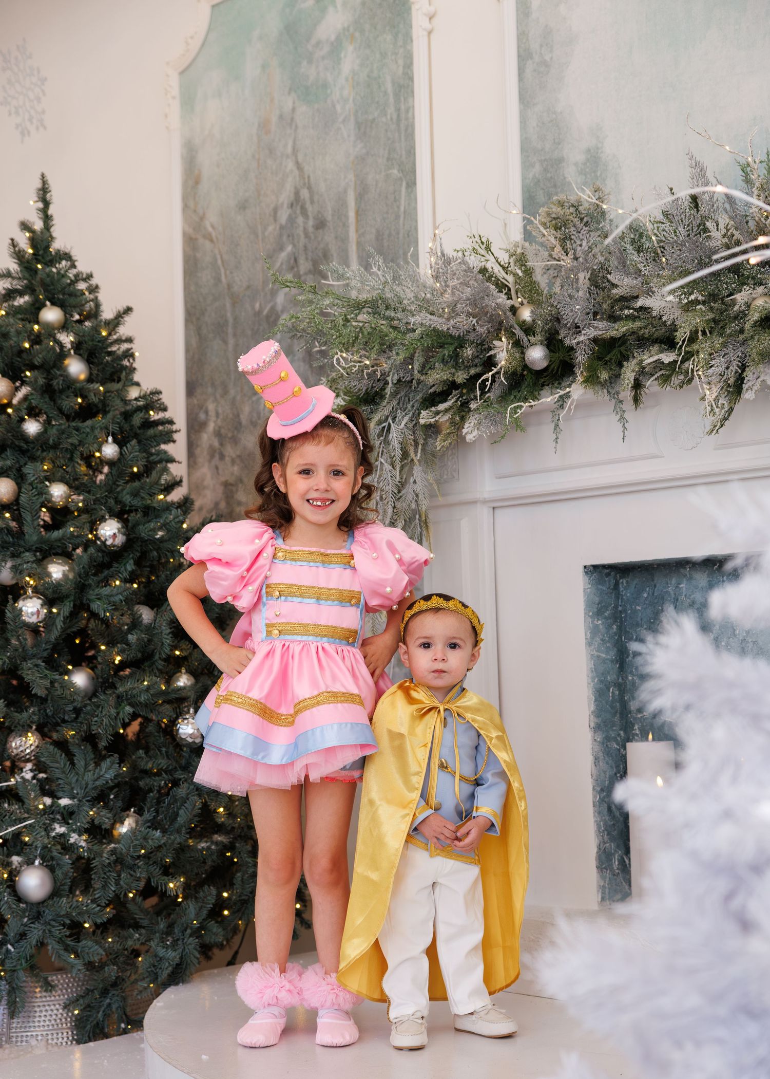 Two toddlers in festive holiday outfits pose together in front of a Christmas tree and decorative garland.