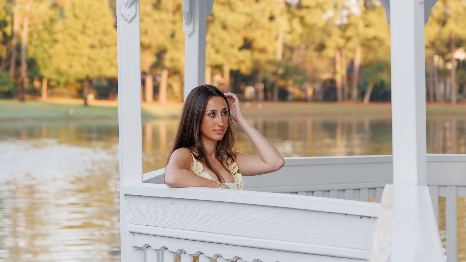 Woman with long dark hair leaning on a white gazebo railing beside a calm lake, with golden autumn trees in the background.