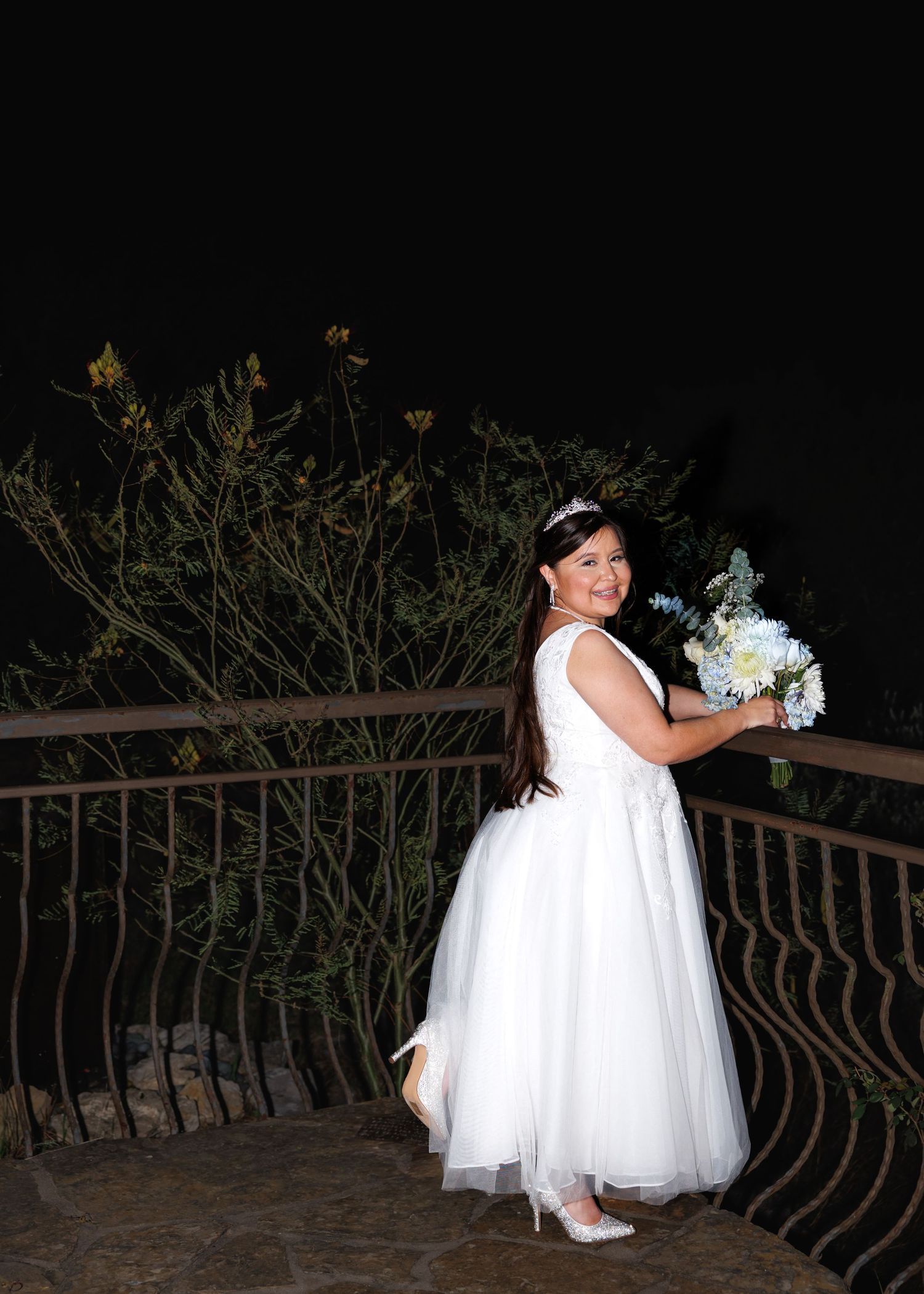 Bride in a white wedding dress poses on a dark outdoor balcony with ornate railings during nighttime photography.