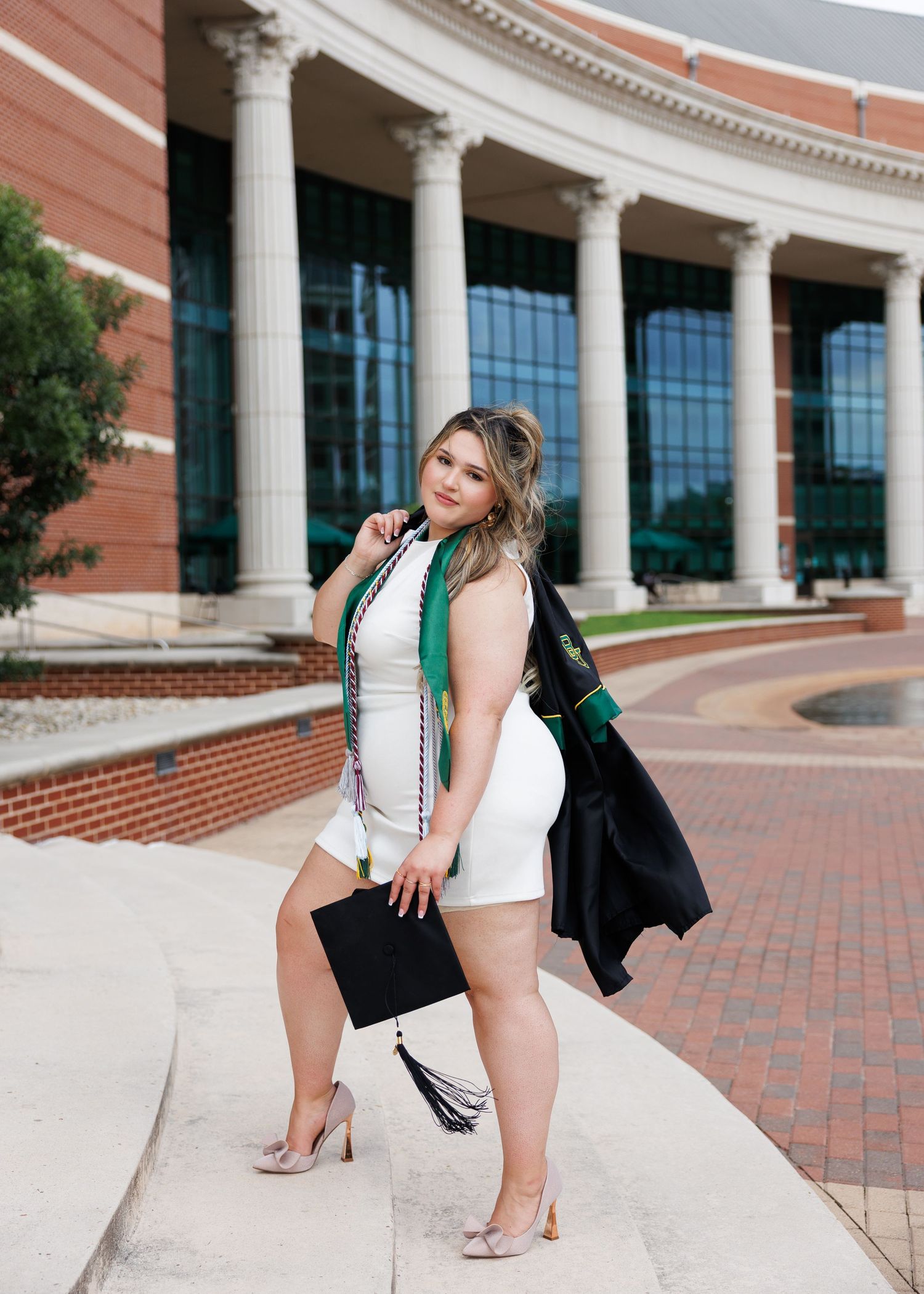 Graduate in a white dress and emerald stole poses in front of a grand columned university building with brick facade.
