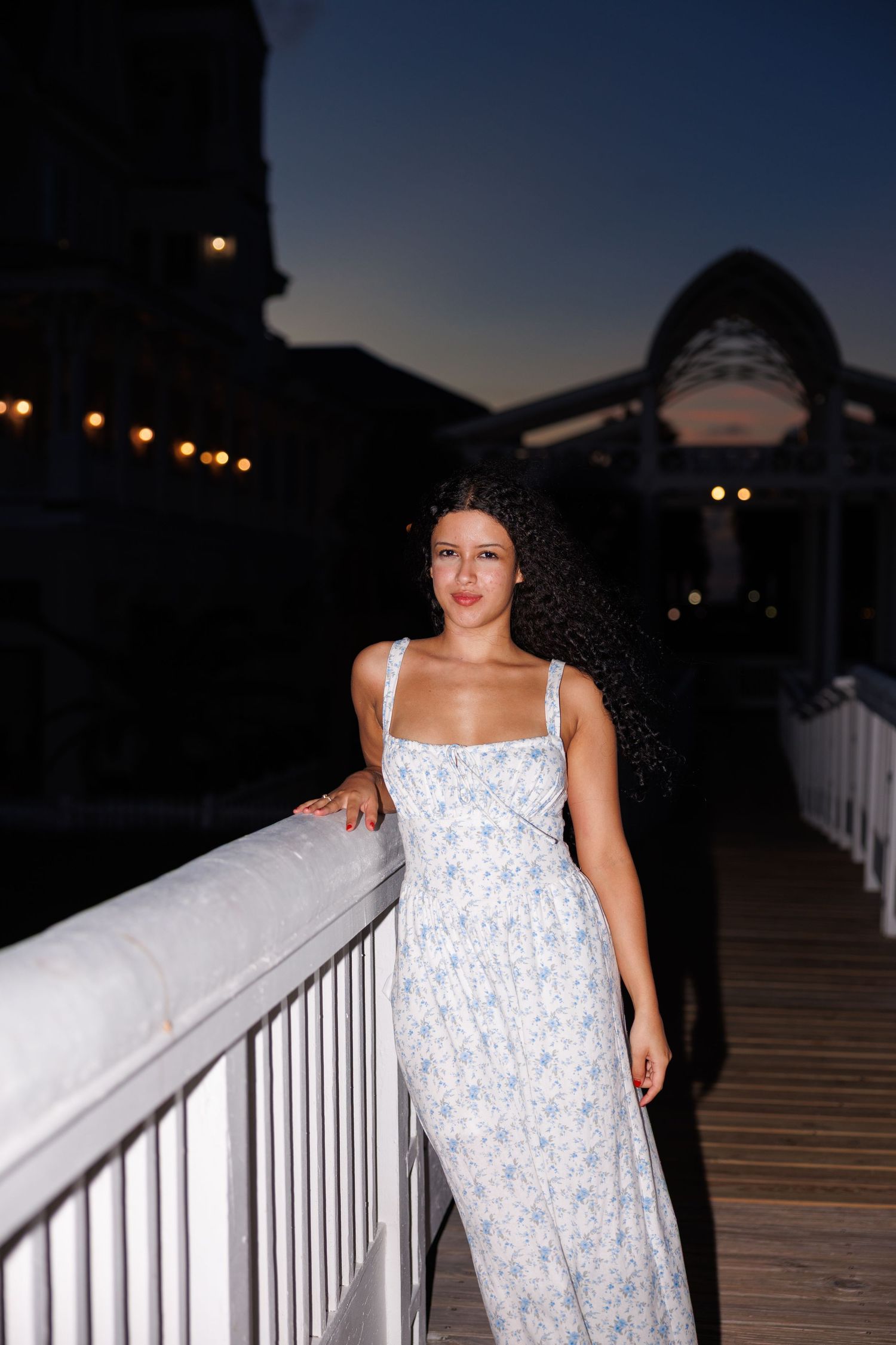 A woman in a flowing white dress stands on an illuminated boardwalk at dusk against a darkening sky.