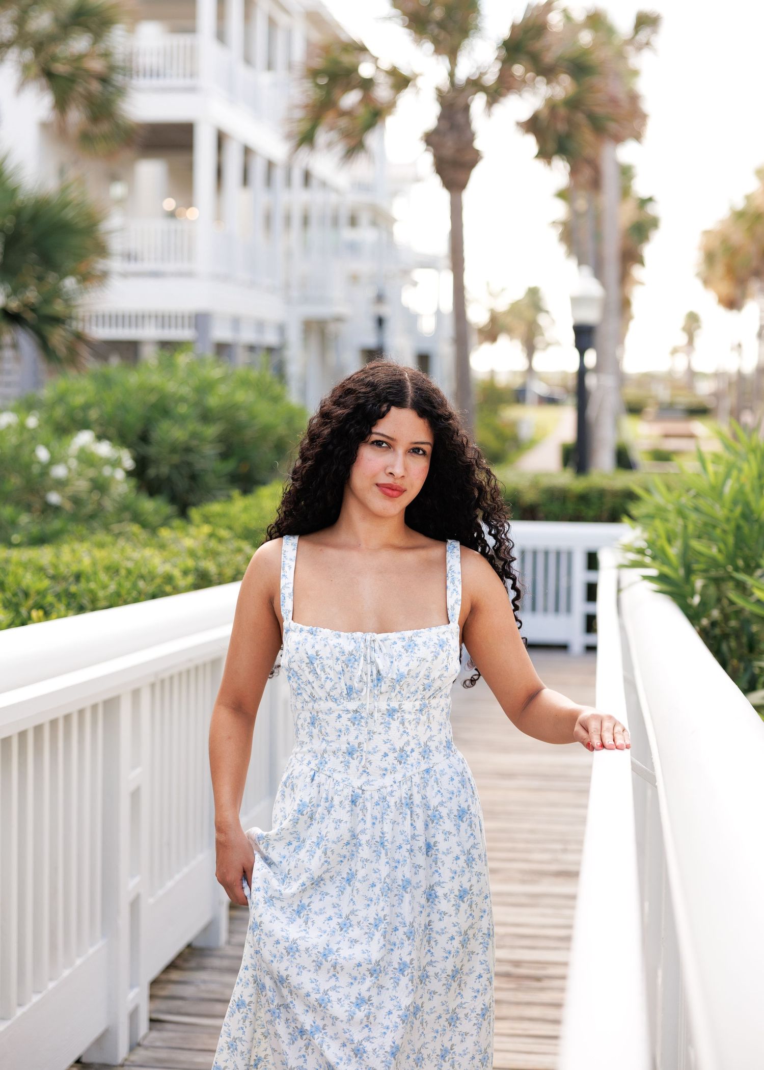 A woman in a blue and white floral sundress poses on a white boardwalk railing in an outdoor beachside setting.