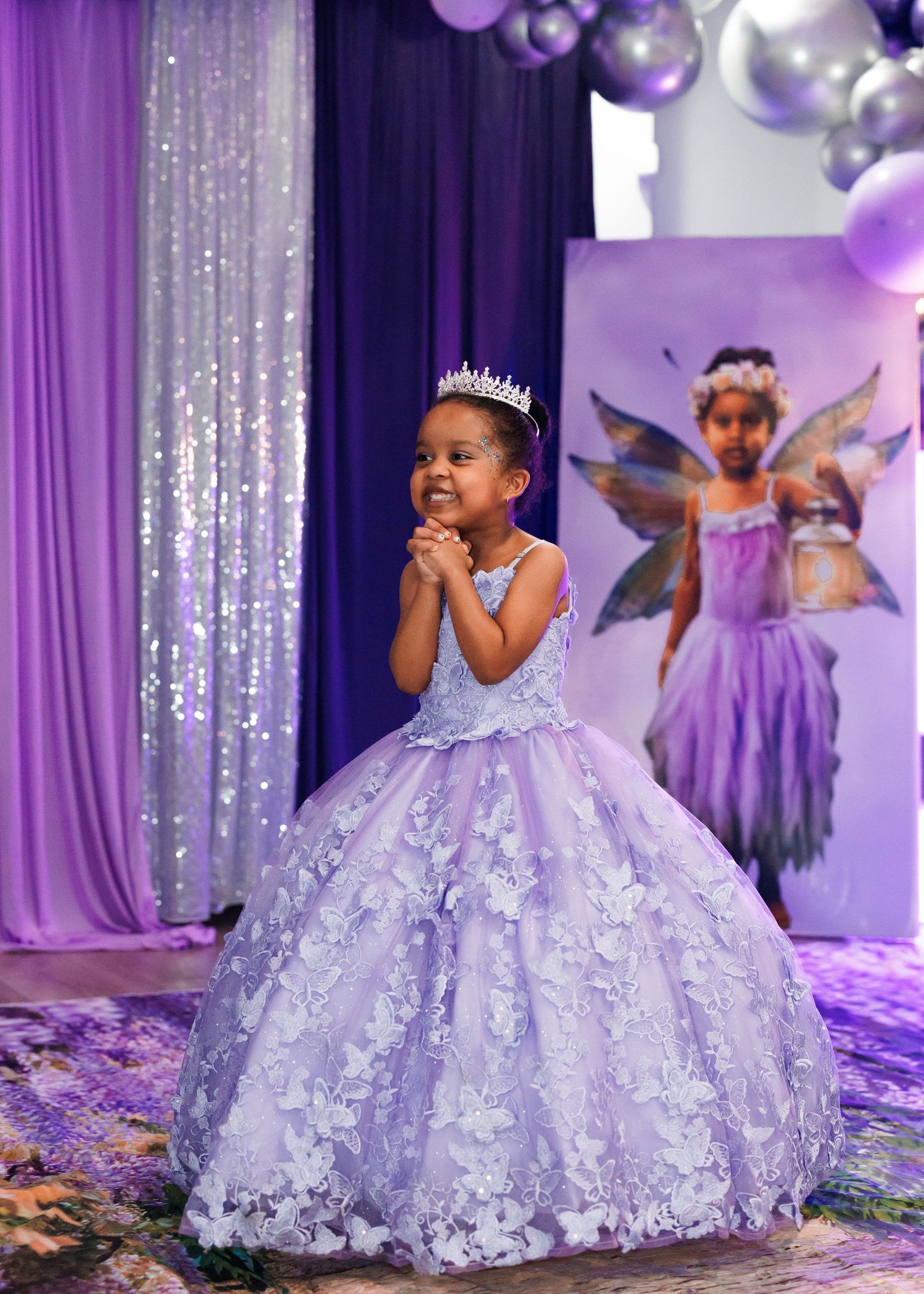 Young girl in lavender ball gown with floral appliques and tiara poses at fairy-themed birthday party.