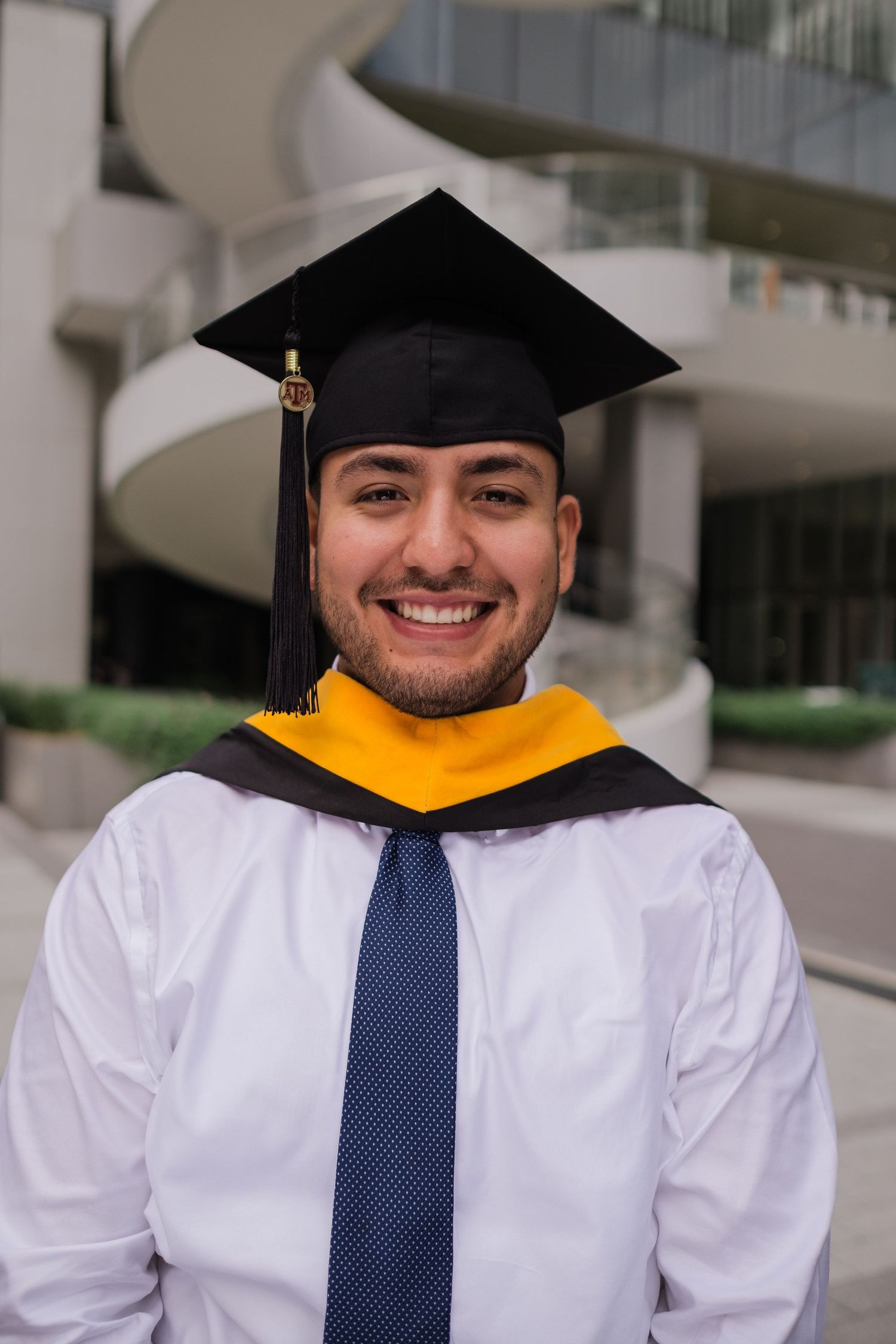 Graduate in white dress shirt and gold hood wearing mortarboard cap and navy tie smiles outdoors.