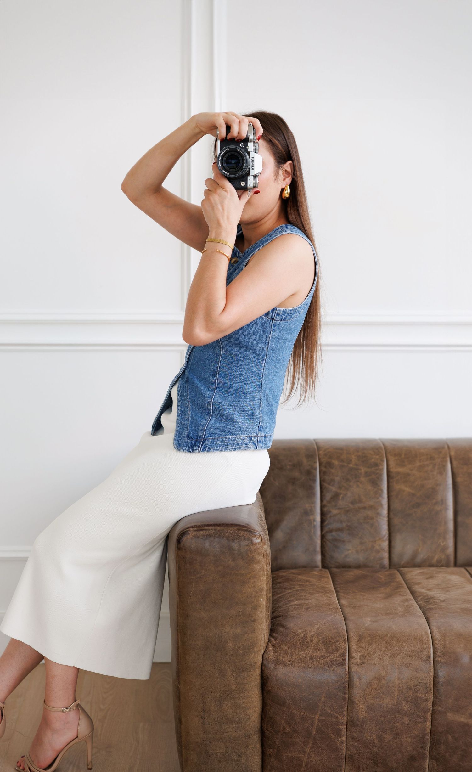 Person in denim top and white pants taking a photo while sitting on brown leather couch.