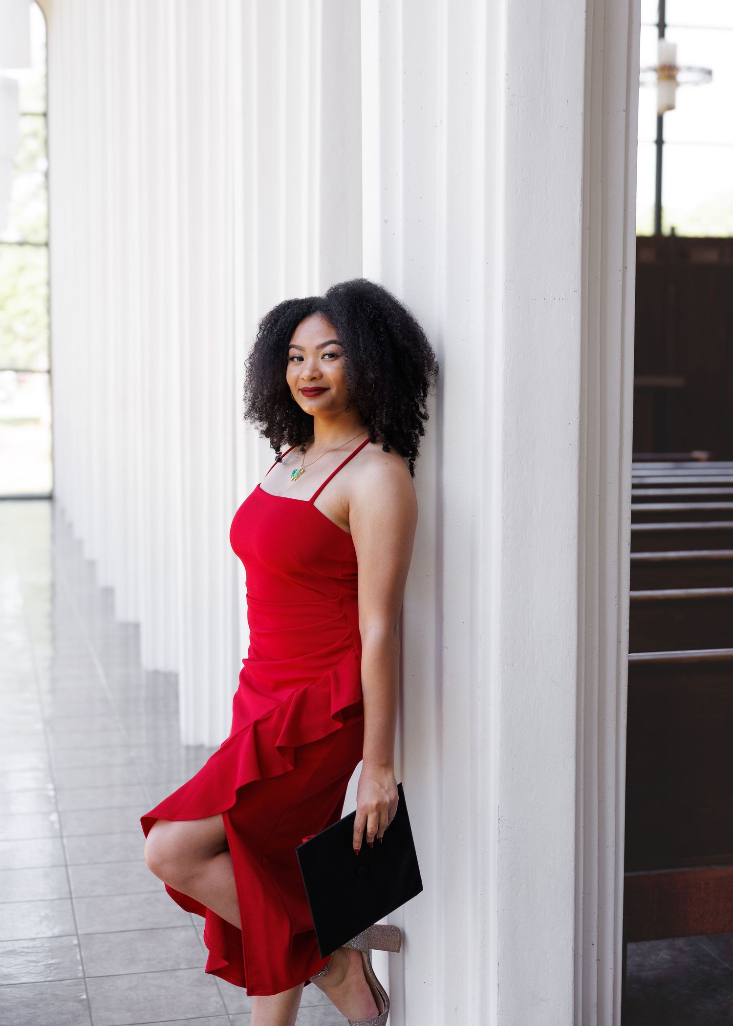 Graduate in a red dress poses elegantly between white columns during graduation photo session.