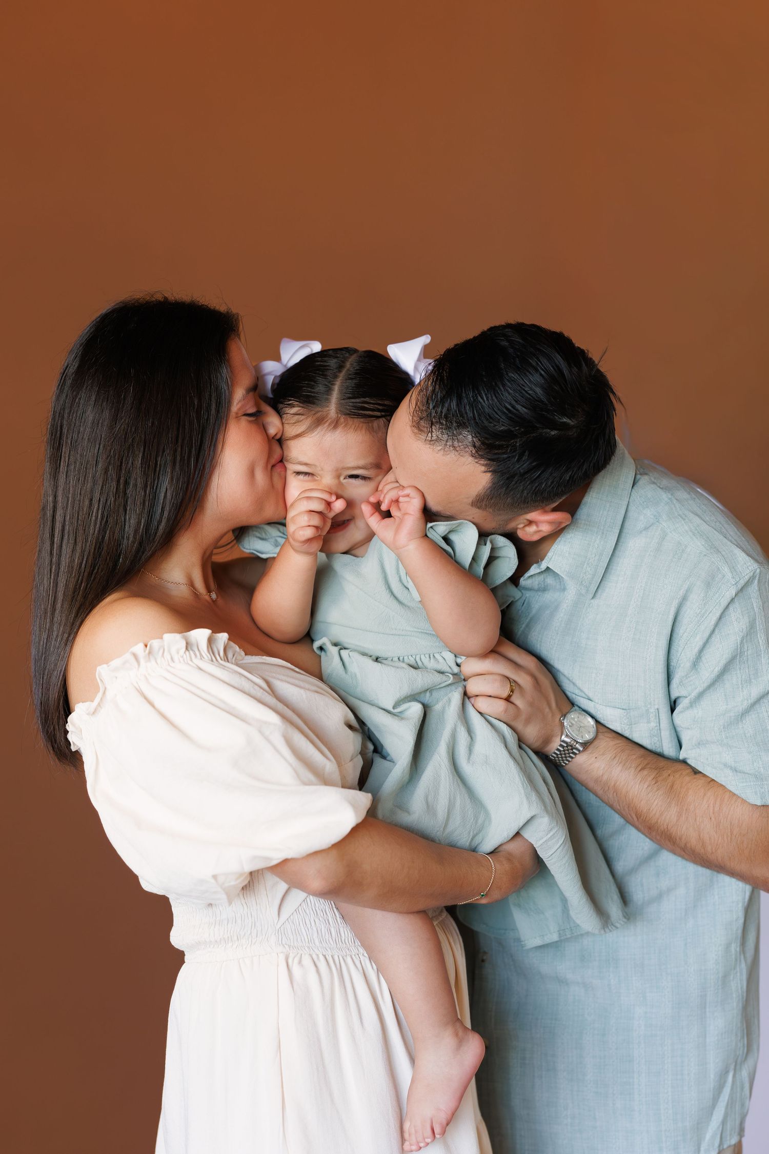 Parents kissing their smiling toddler daughter on both cheeks against a warm brown studio background.