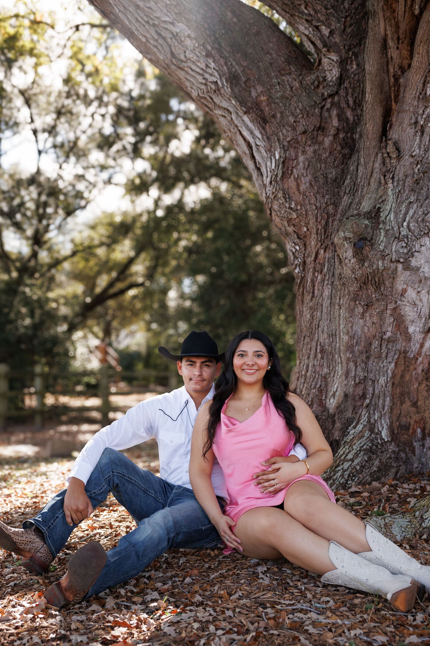 A young couple sits together under a large tree, man in cowboy hat and woman in pink dress, smiling outdoors.