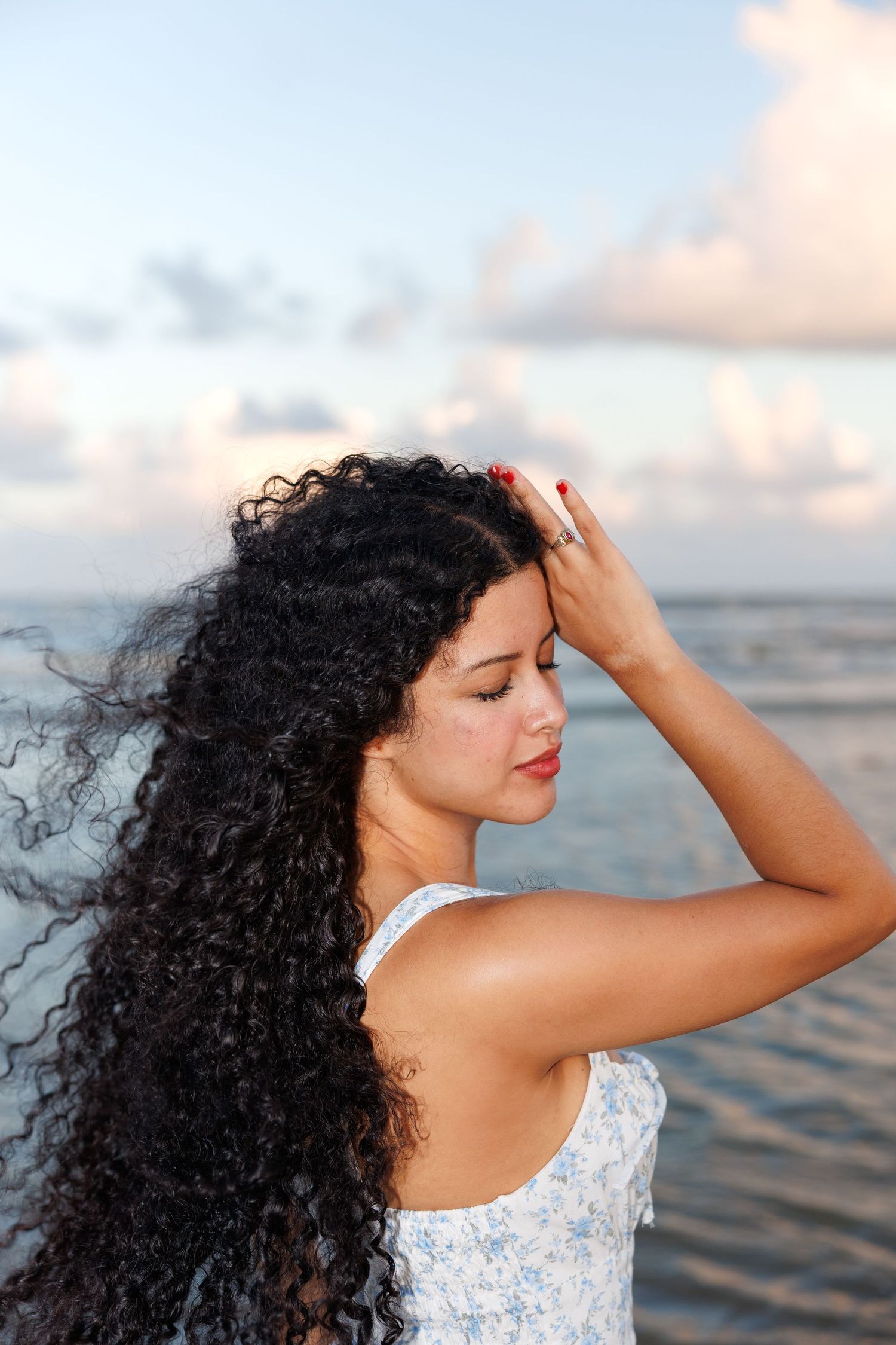 Long dark curly hair blows in the breeze as woman in white dress poses against sunset ocean backdrop.