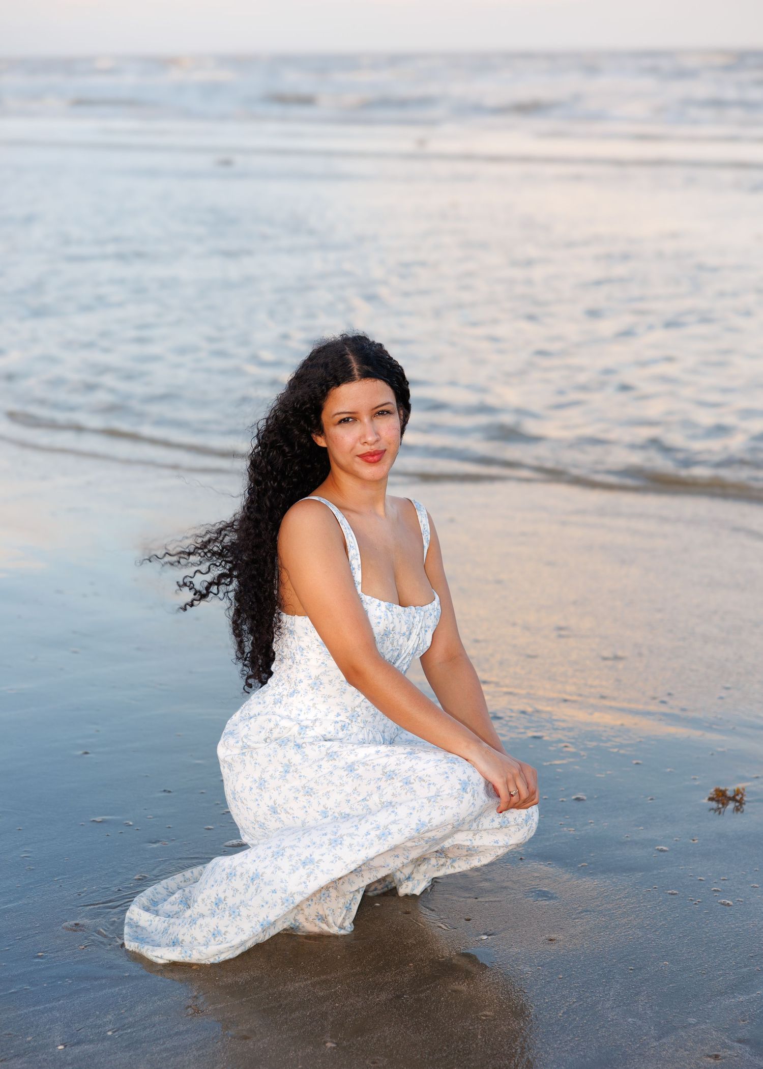 A person in a flowing white dress sits on a beach at sunset.