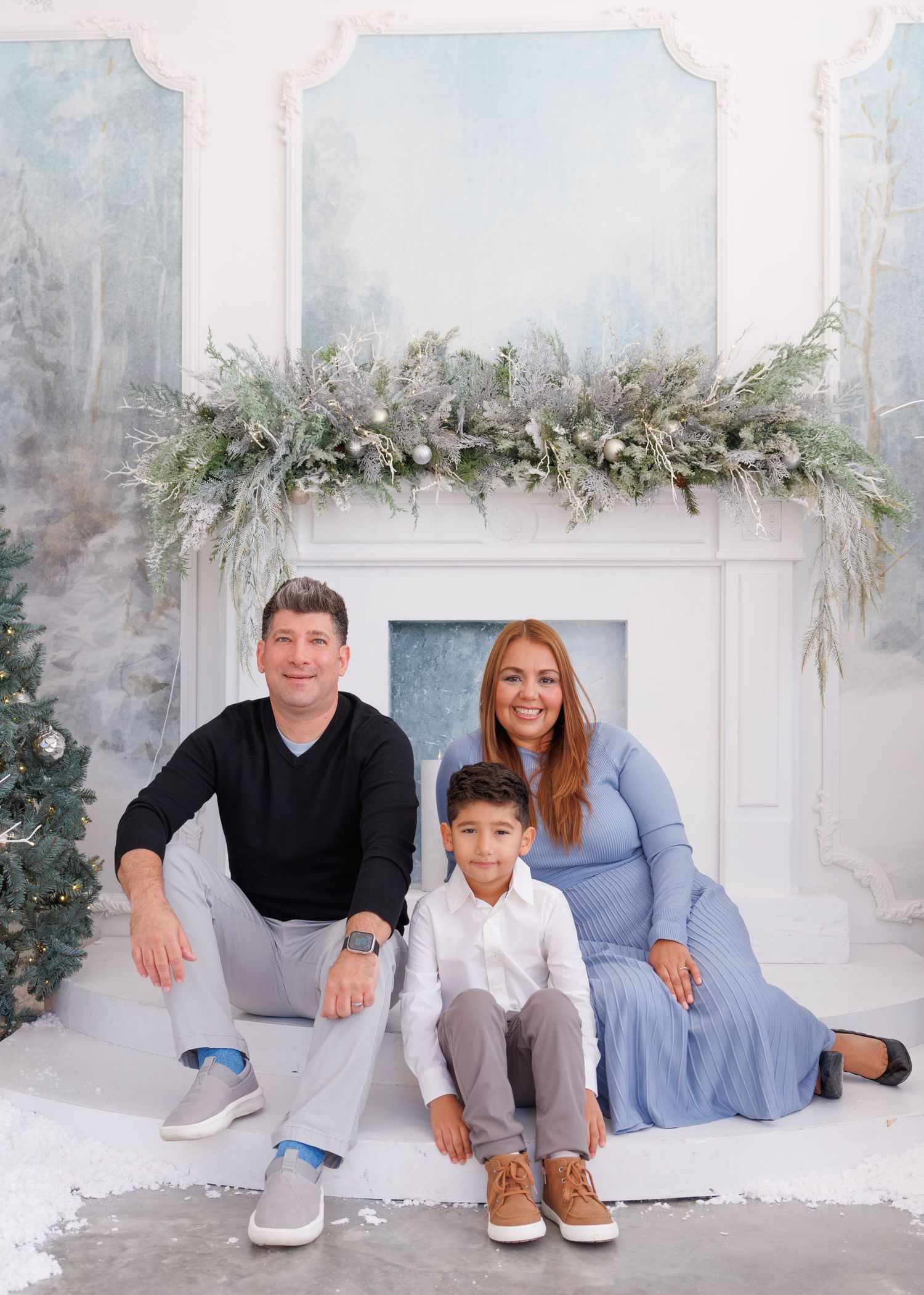 A family poses together in front of a white fireplace mantel decorated with elegant winter greenery and garland.