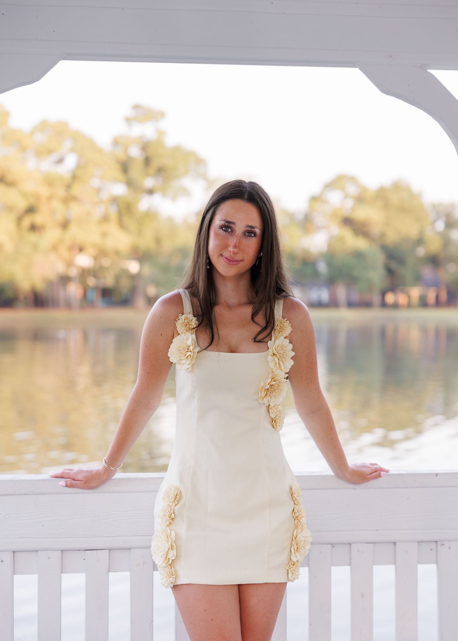 Woman with long brown hair in a cream mini dress with floral appliqué details leaning on a white railing by a calm lake at sunset.