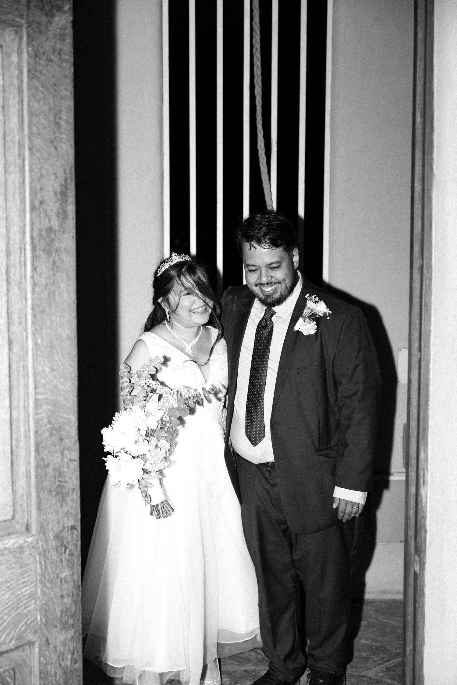 A black and white photo of a happy couple at their wedding, the bride in white and groom in a suit with boutonniere.