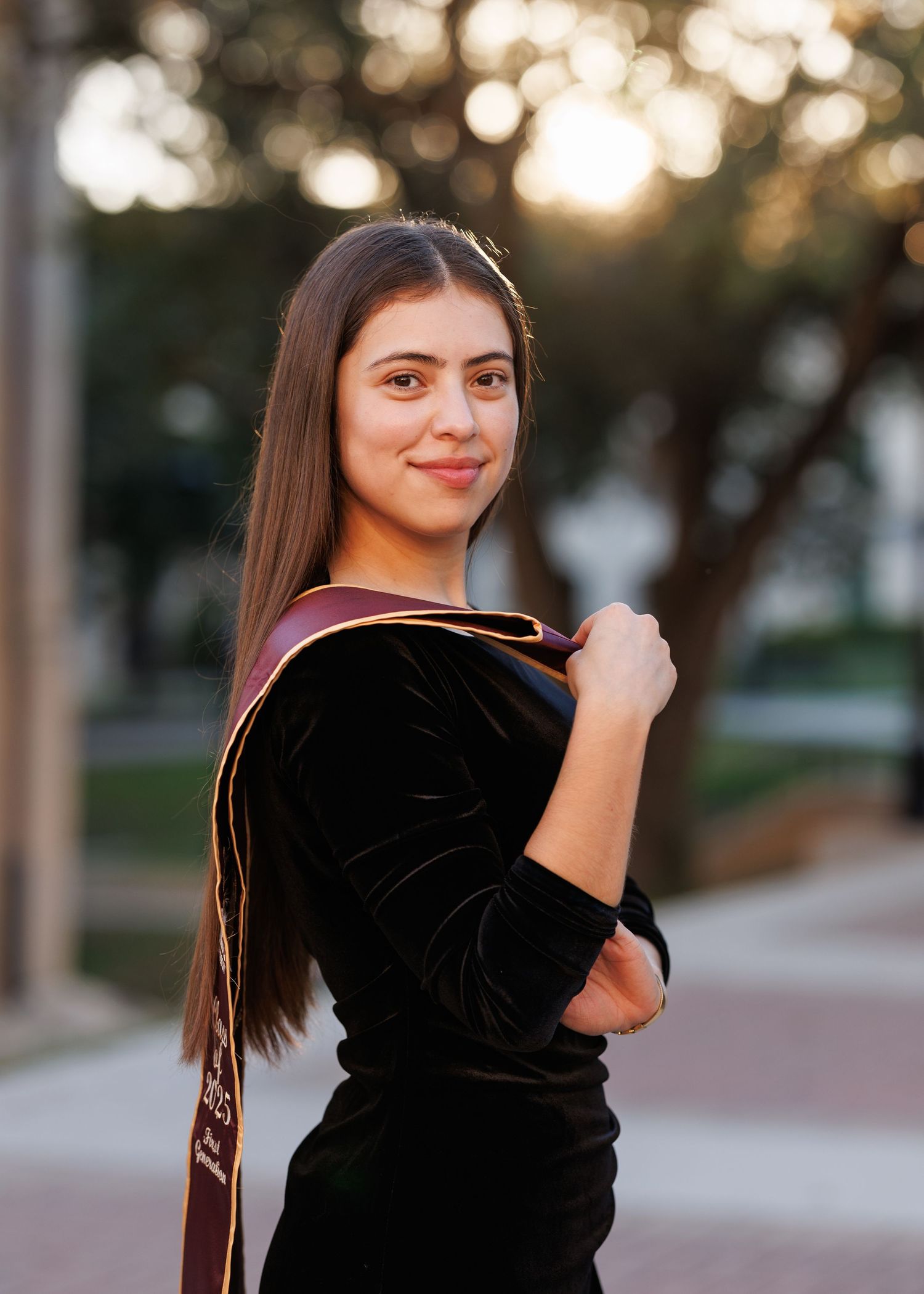 Person in black velvet top poses outdoors at sunset with soft bokeh lighting in background.