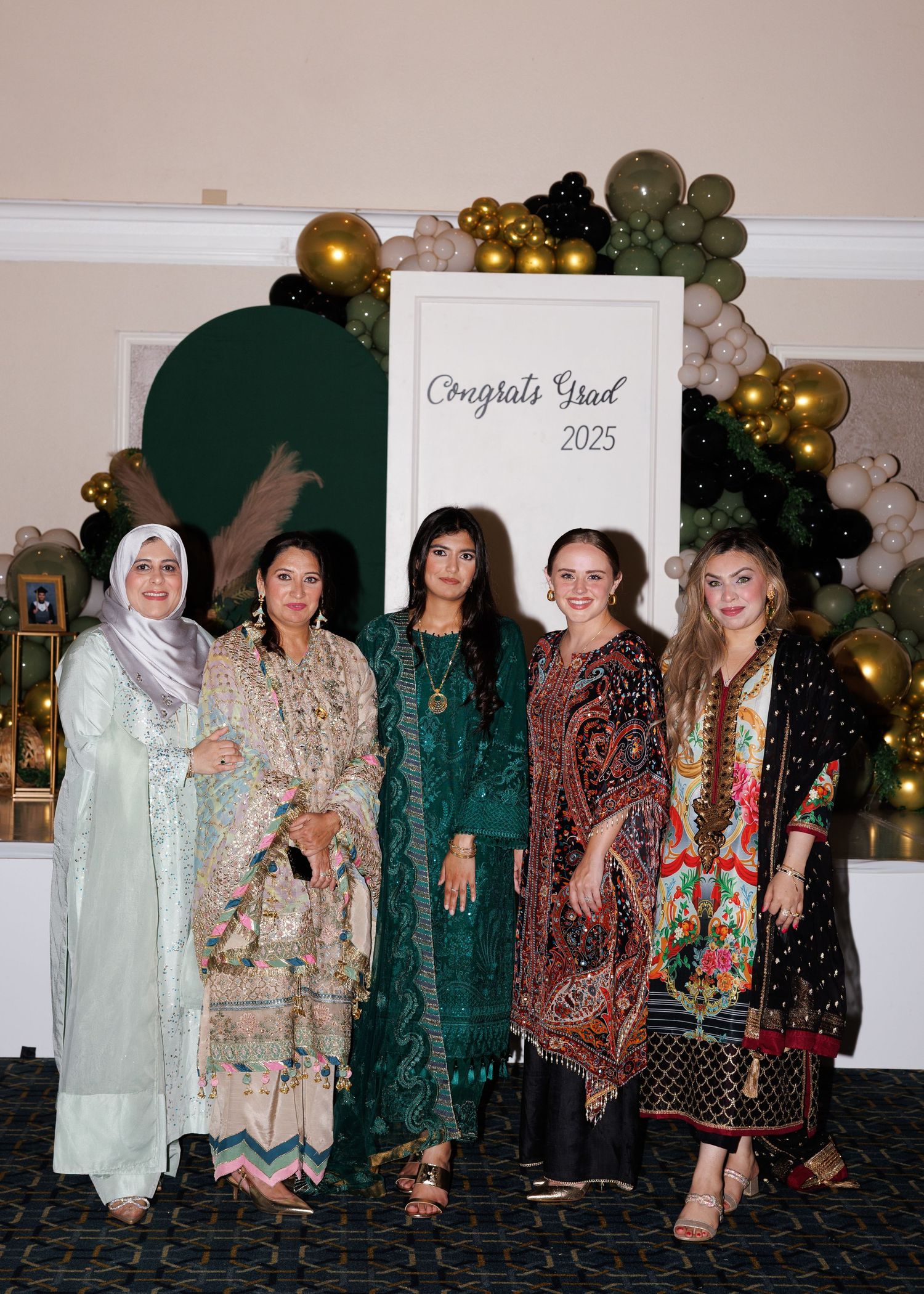 A group of five women in formal traditional attire posing at a graduation celebration with gold and green balloon decor.