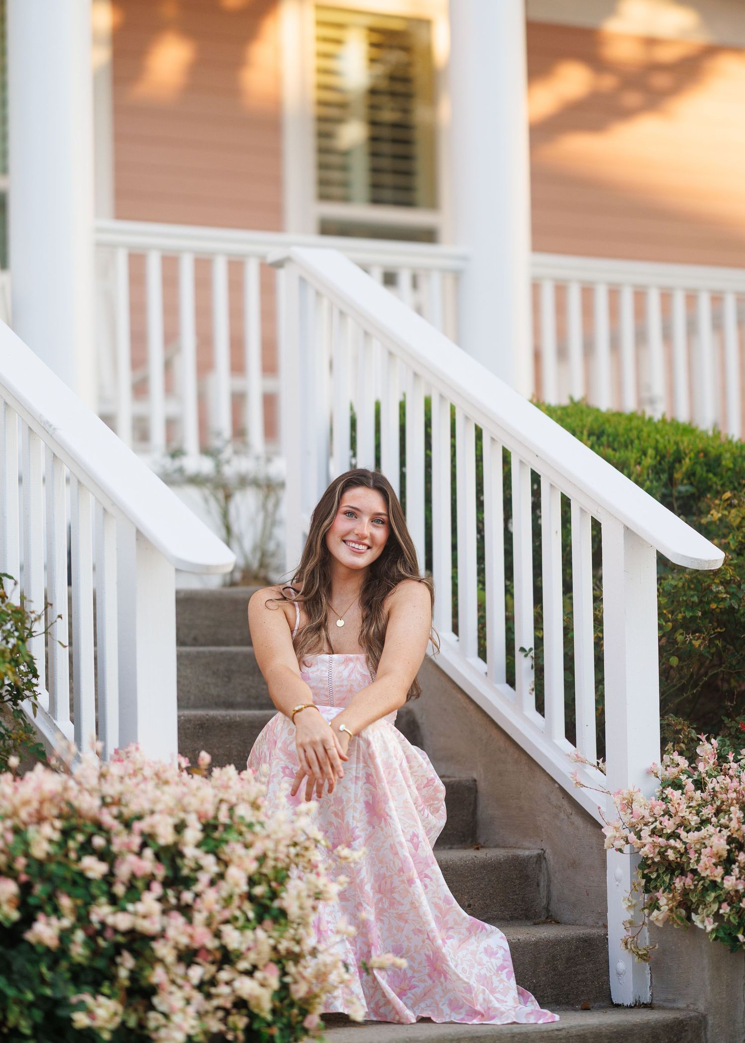 Smiling young woman in a pink floral dress sits on porch steps beside white railings and blooming pink flowers.