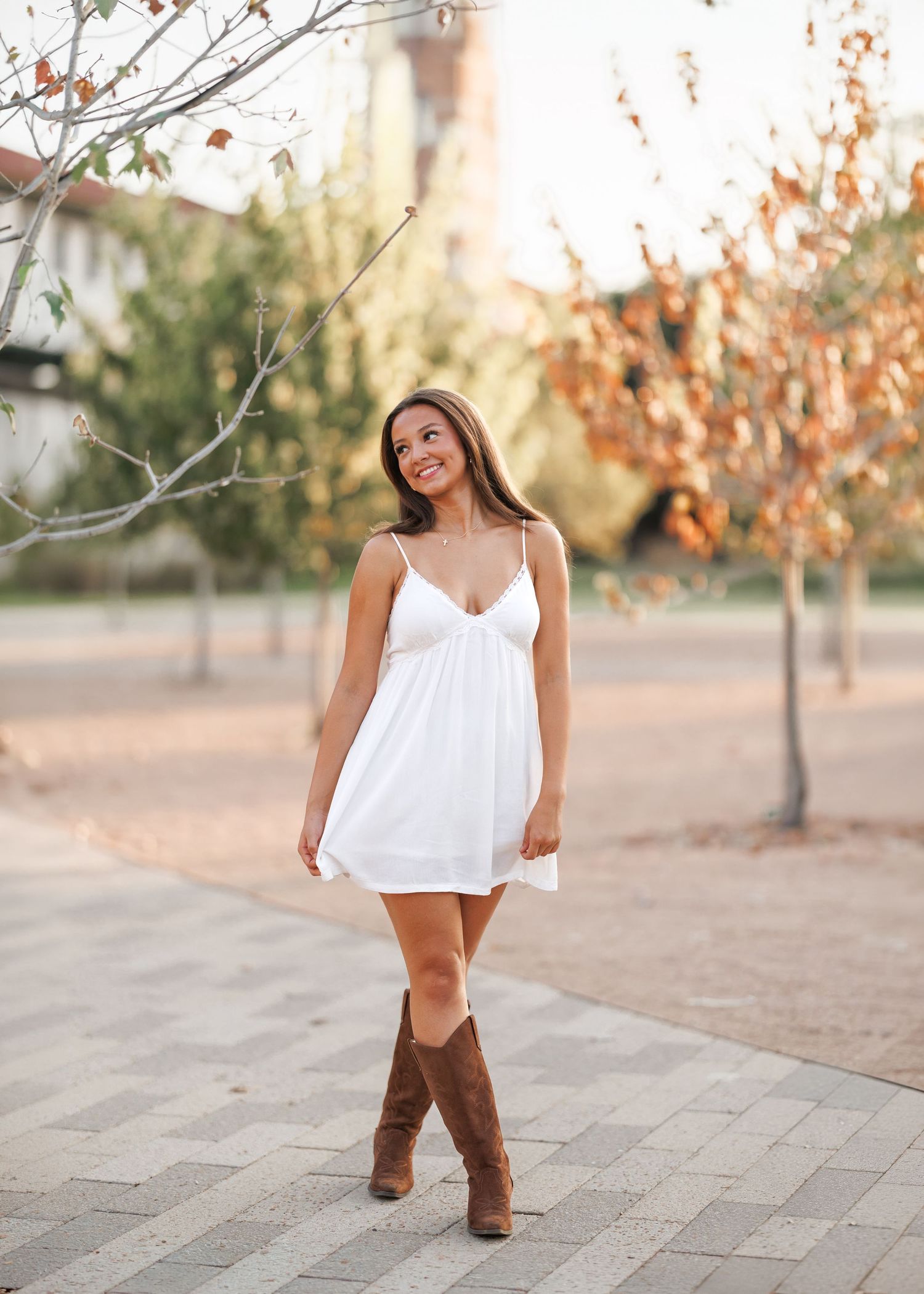 Smiling young woman in a white sundress and brown cowboy boots poses on a brick paved path lined with autumn trees.