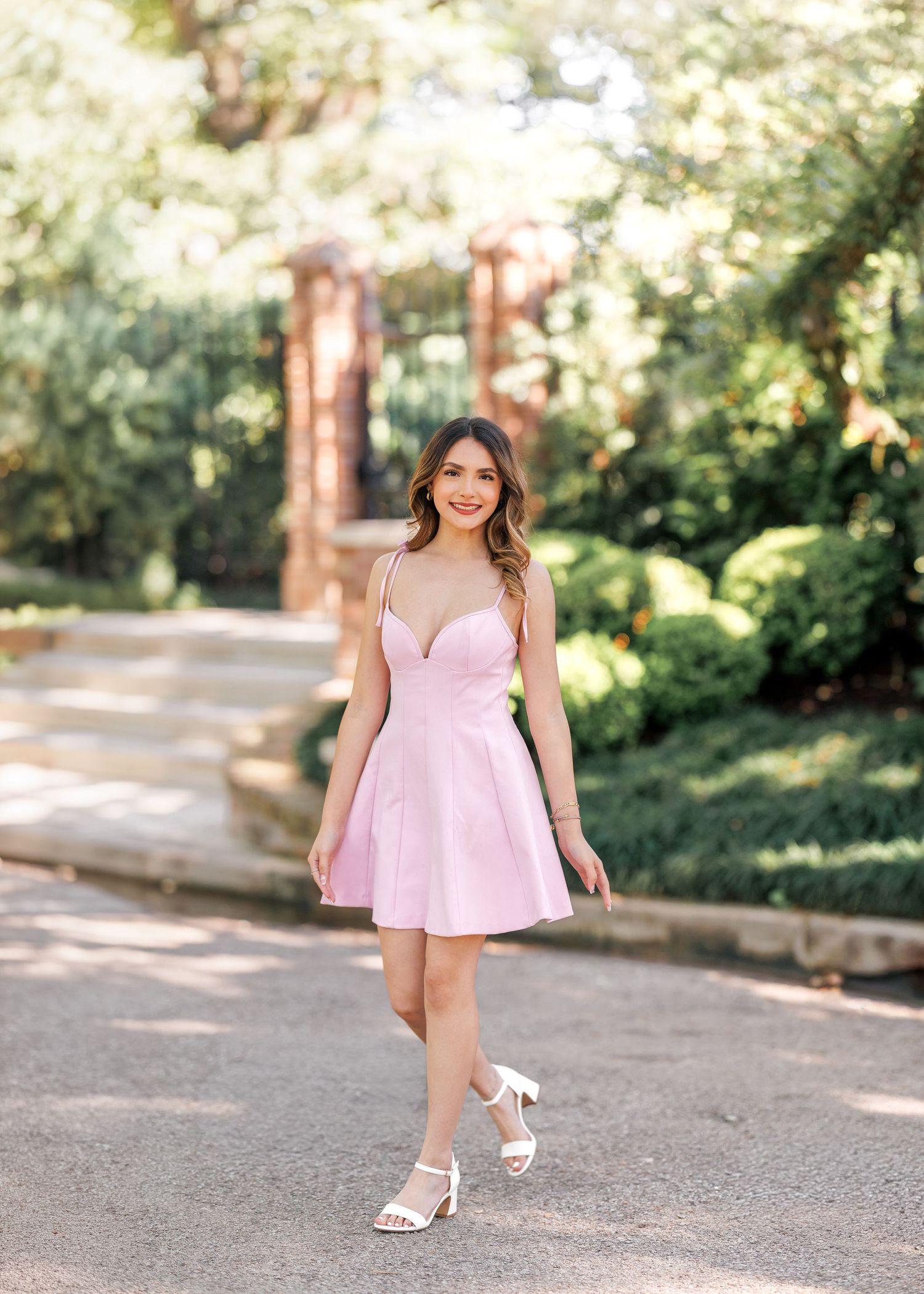 Young woman smiling in a pink mini dress and white heels, posing outdoors in a lush garden setting.