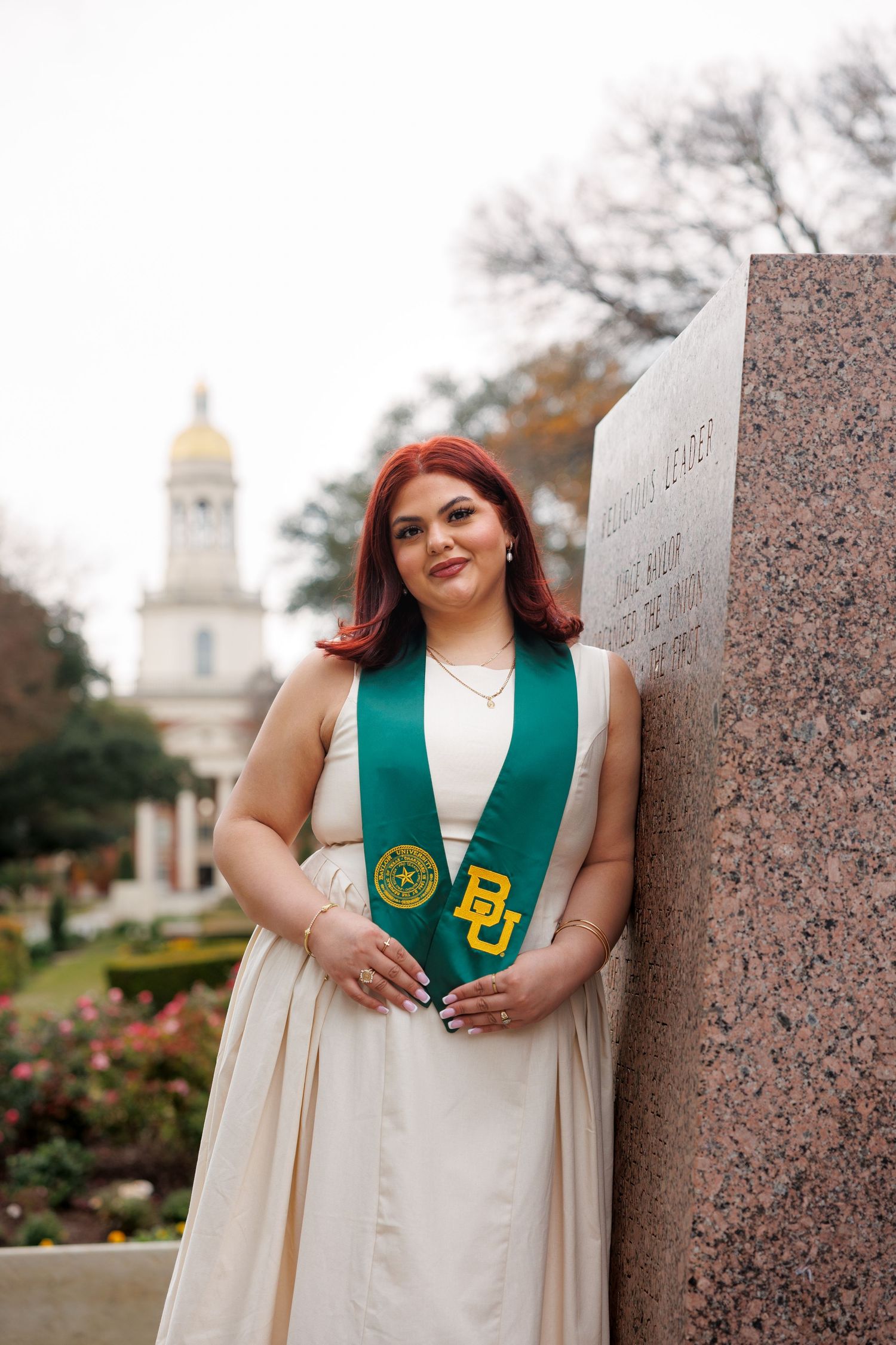 Graduate wearing white dress and green graduation stole poses against pink granite wall with campus building in background.