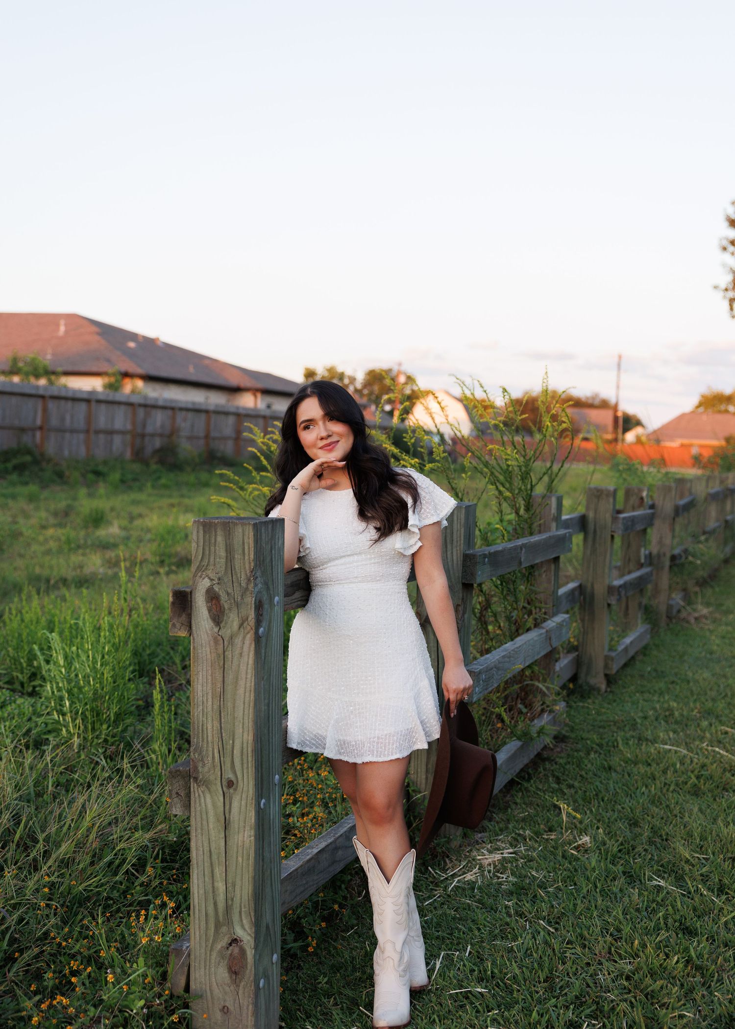 Woman in white dress poses along rustic wooden fence in countryside setting during sunset.