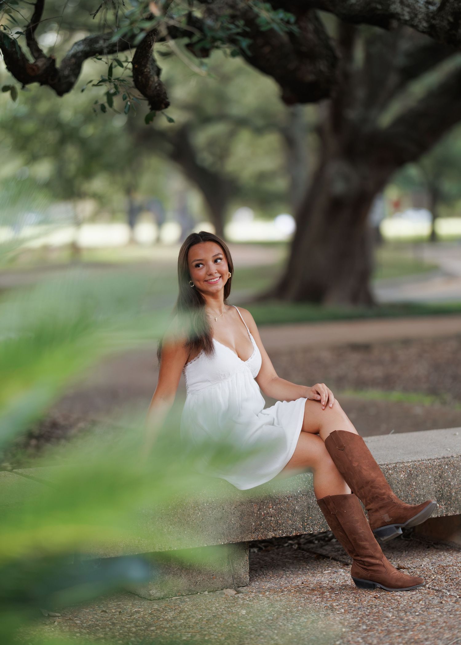 Smiling young woman in a white sundress and brown cowboy boots sits on a stone bench beneath large oak trees in a sunny park.