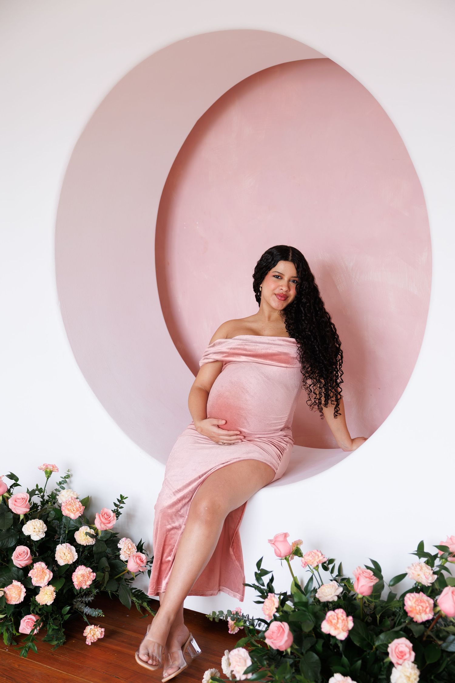 Pregnant woman in pink off-shoulder dress sits in circular pink alcove surrounded by roses, cradling her baby bump.