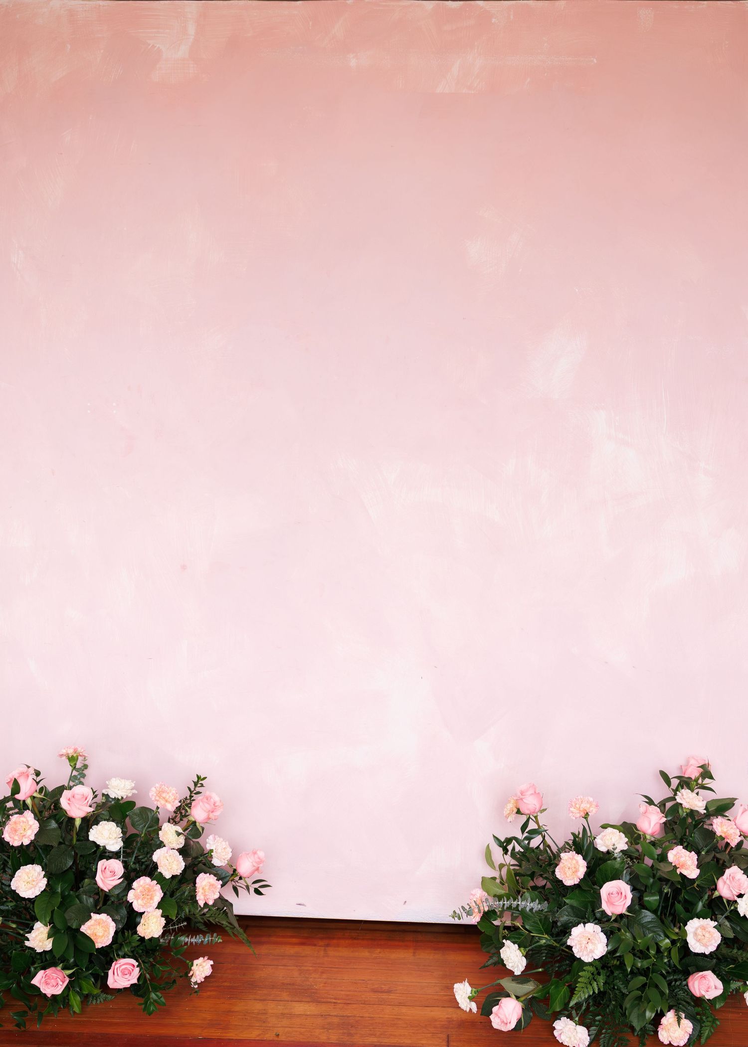 Pink roses and green foliage arranged along a wooden ledge against a soft pink textured wall backdrop.