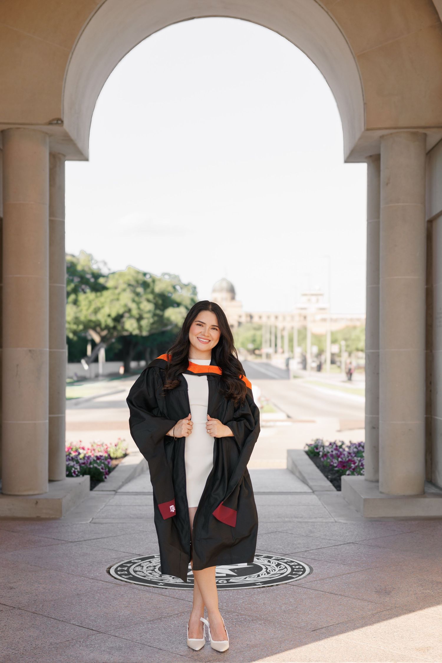 Smiling graduate in black gown with orange trim poses under stone arch at university campus.