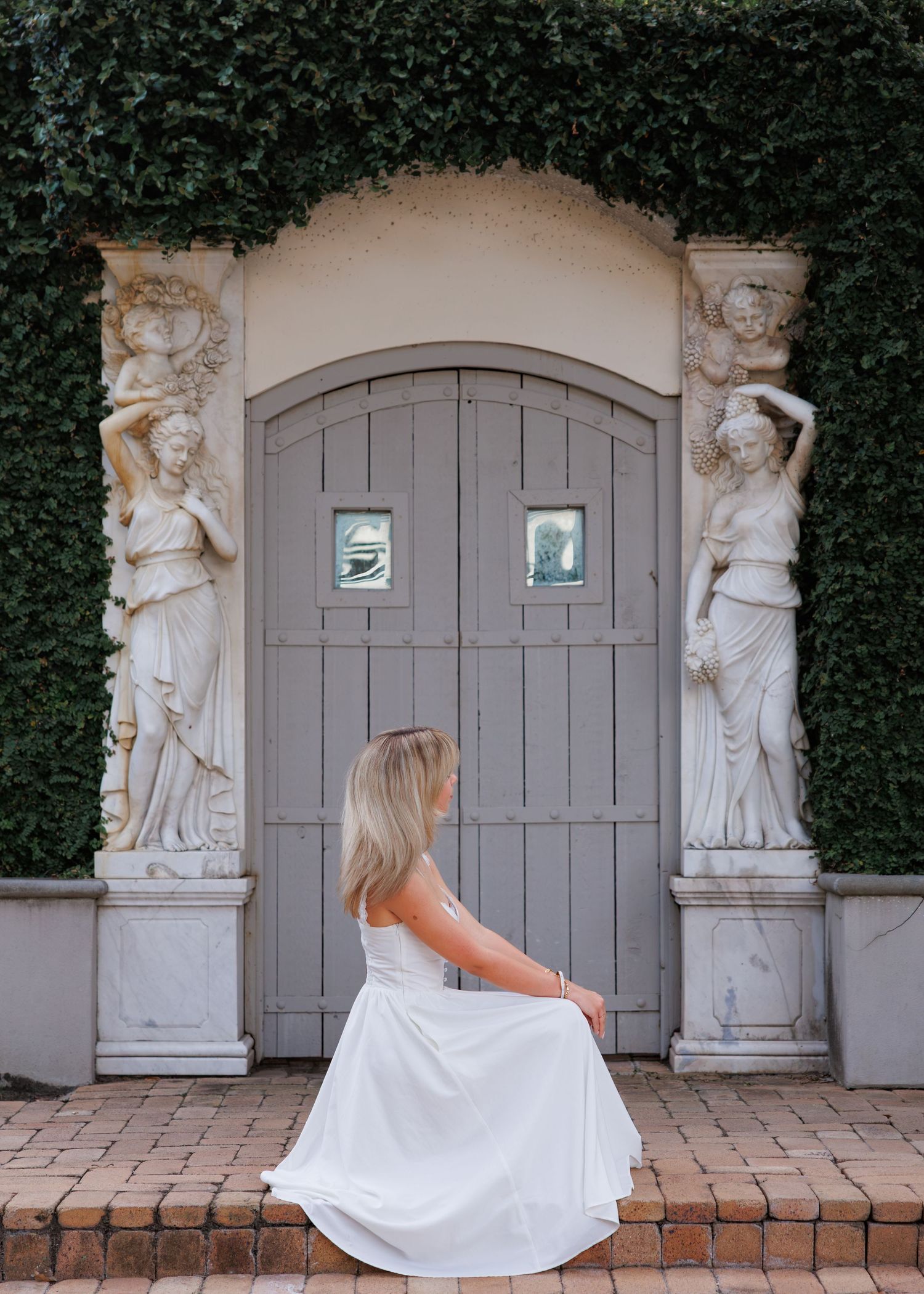 Woman in a flowing white dress sits on brick steps before a gray arched wooden door flanked by stone statues and ivy-covered walls.