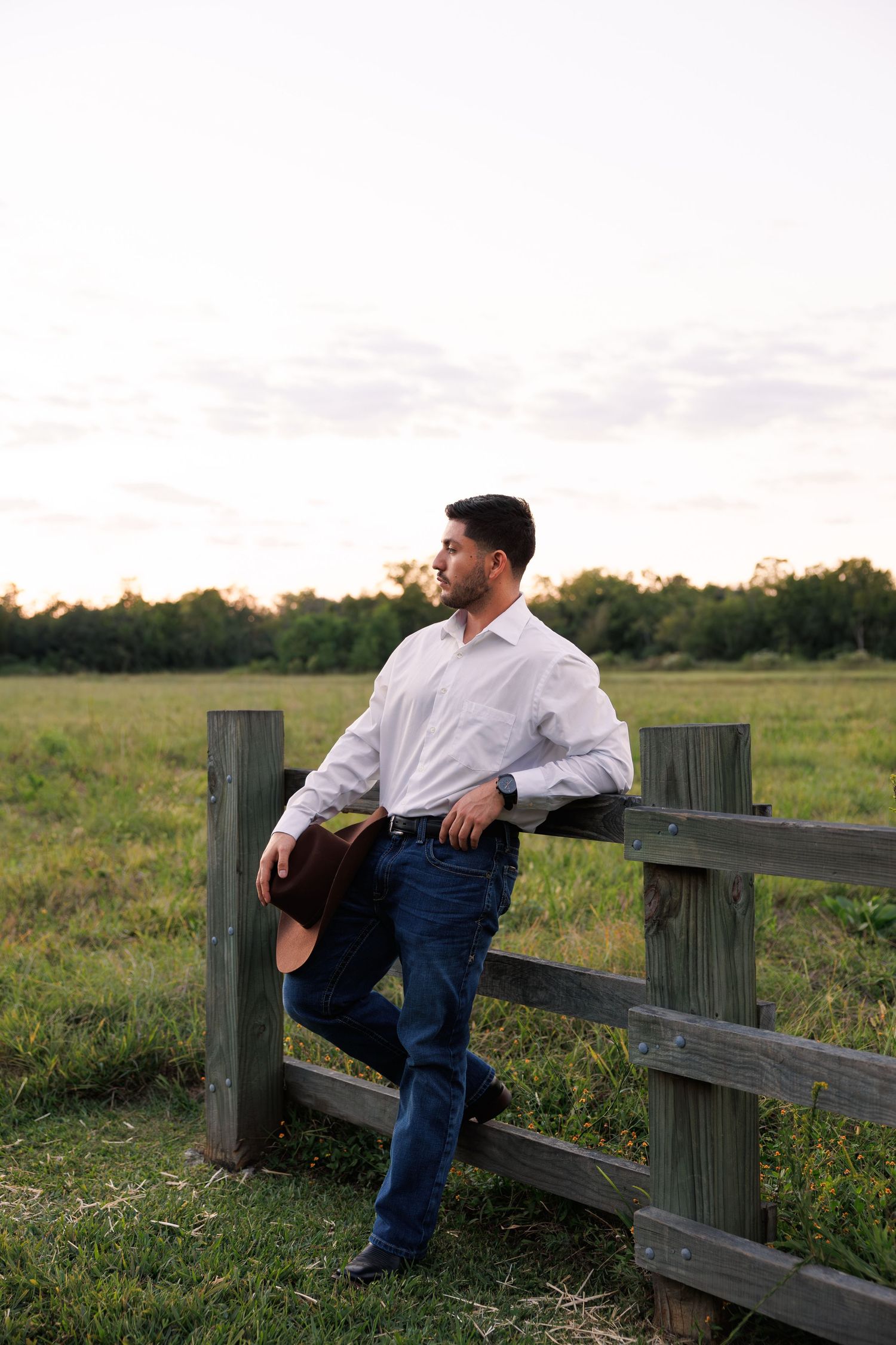 A person in a white shirt and jeans leans against a wooden fence at sunset.