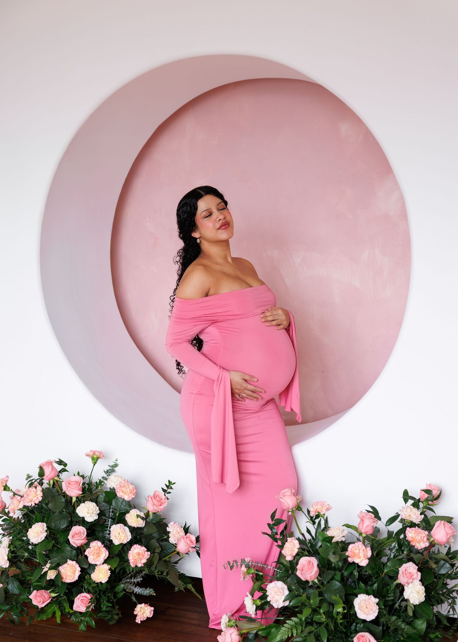 Pregnant woman in pink off-shoulder gown poses gracefully before circular pink backdrop surrounded by roses.
