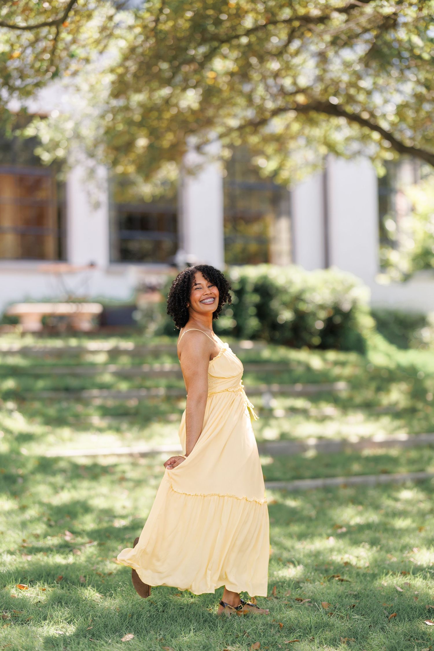 A smiling woman in a flowing yellow maxi dress walks through a lush green park on a sunny day.