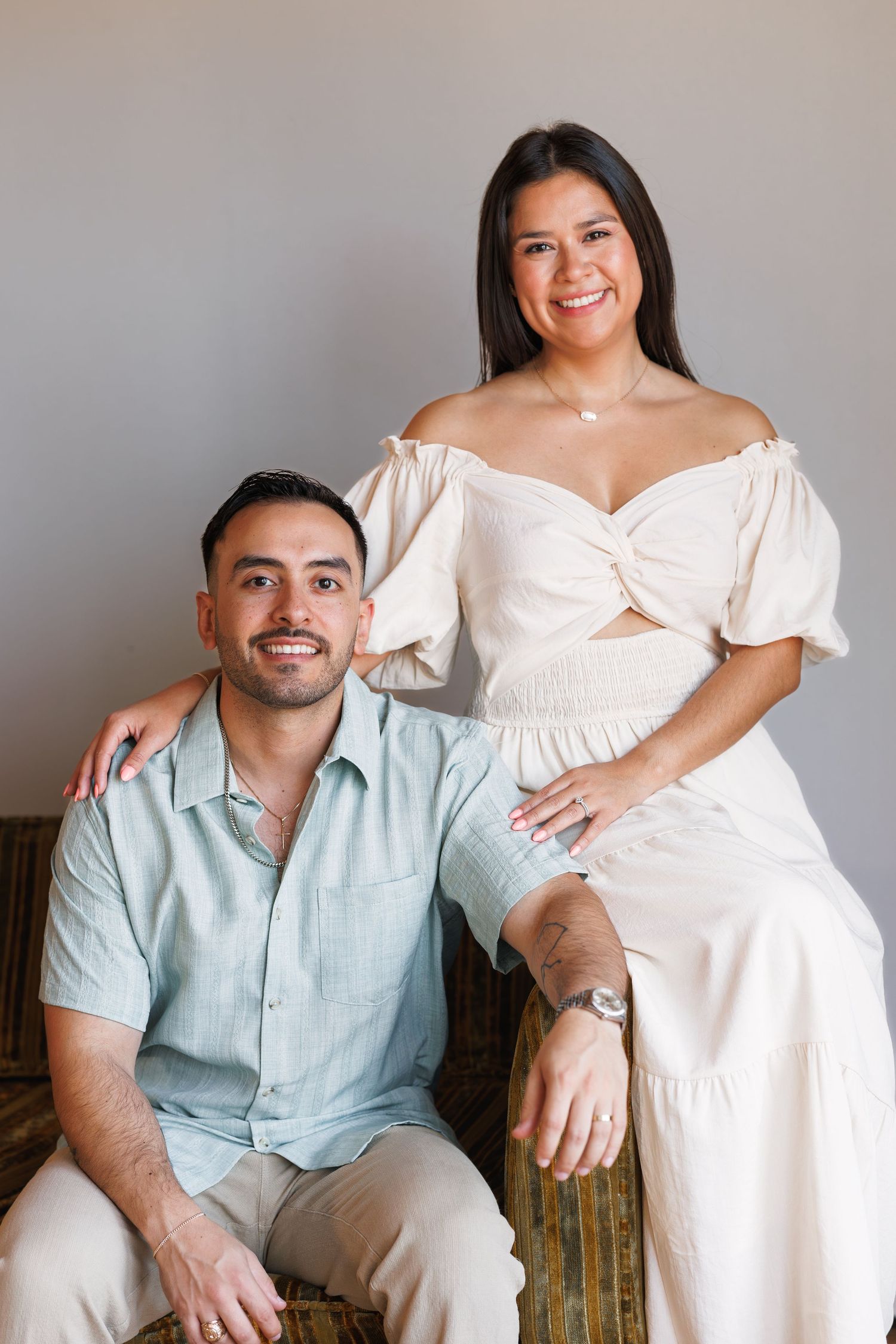 Smiling couple posing together, woman in white off-shoulder dress standing behind seated man in light blue shirt.