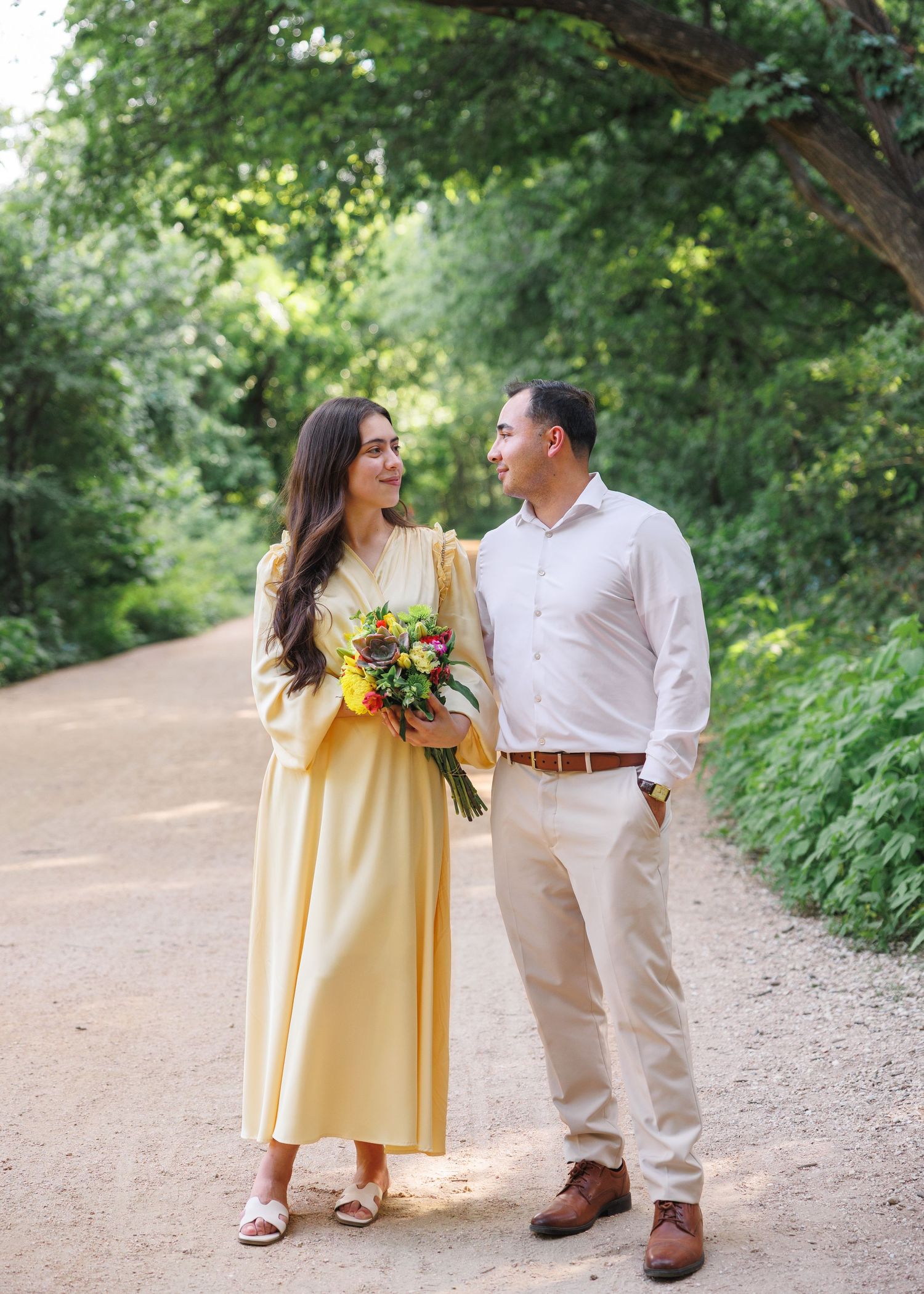 Couple in yellow dress and white shirt holding colorful bouquet on a tree-lined path outdoors.