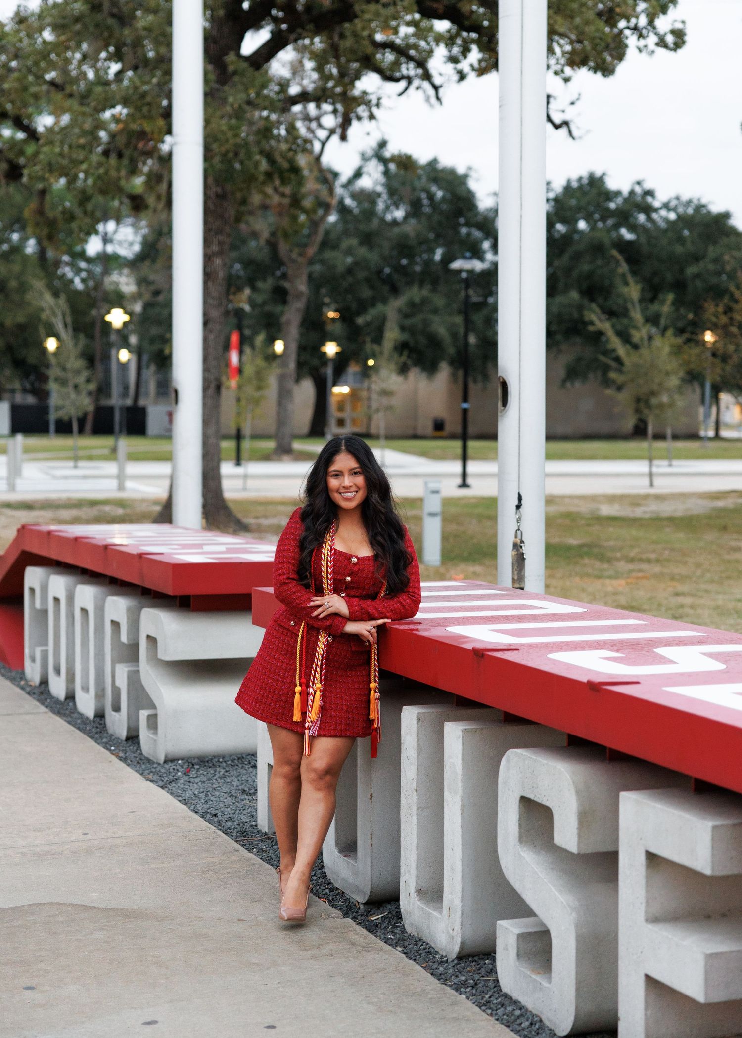 Graduate poses in a red graduation dress next to large white and red block letters at the university campus.
