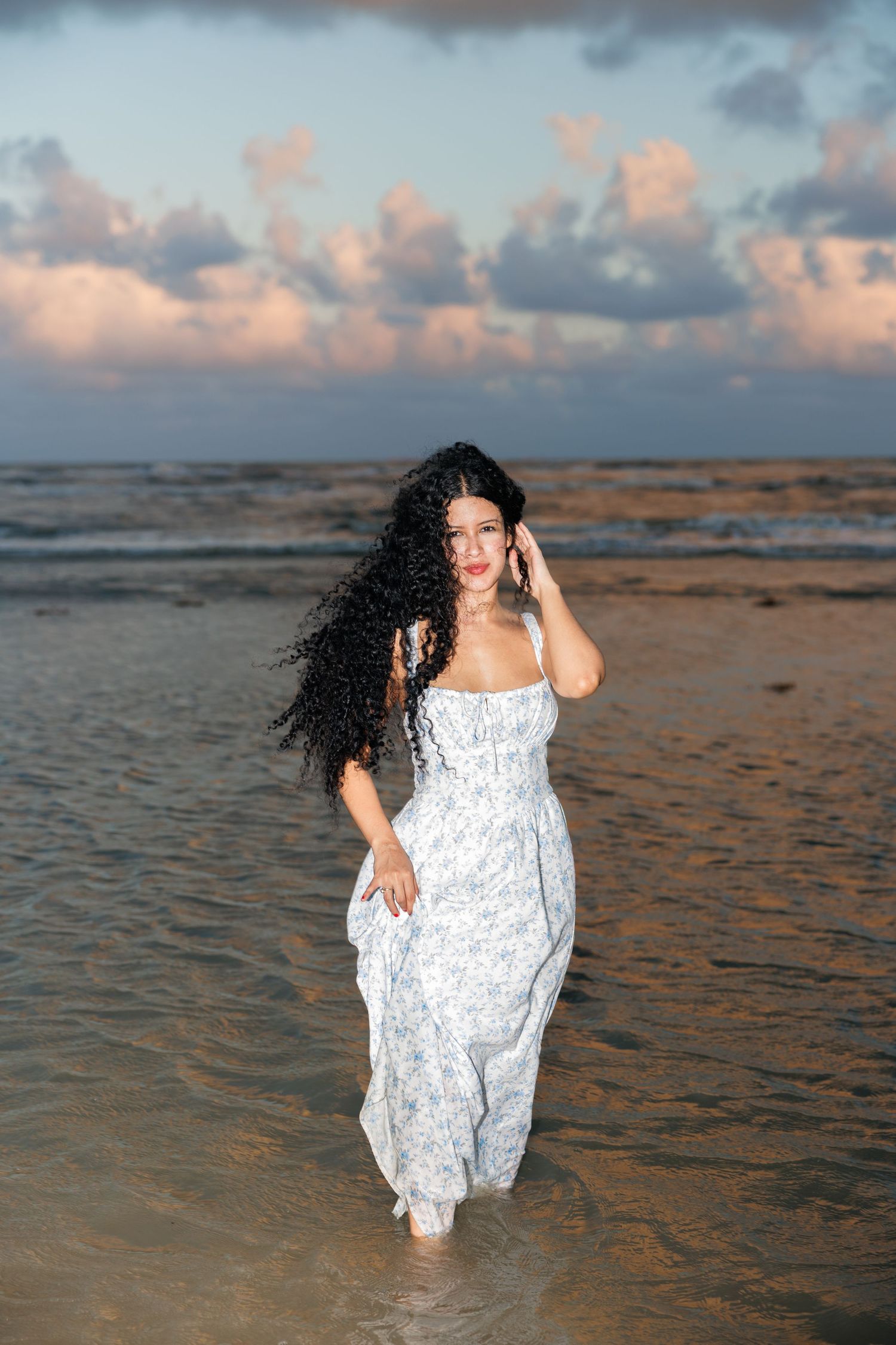A person in a flowing white dress walks barefoot along a beach shoreline during sunset with cloudy skies overhead.