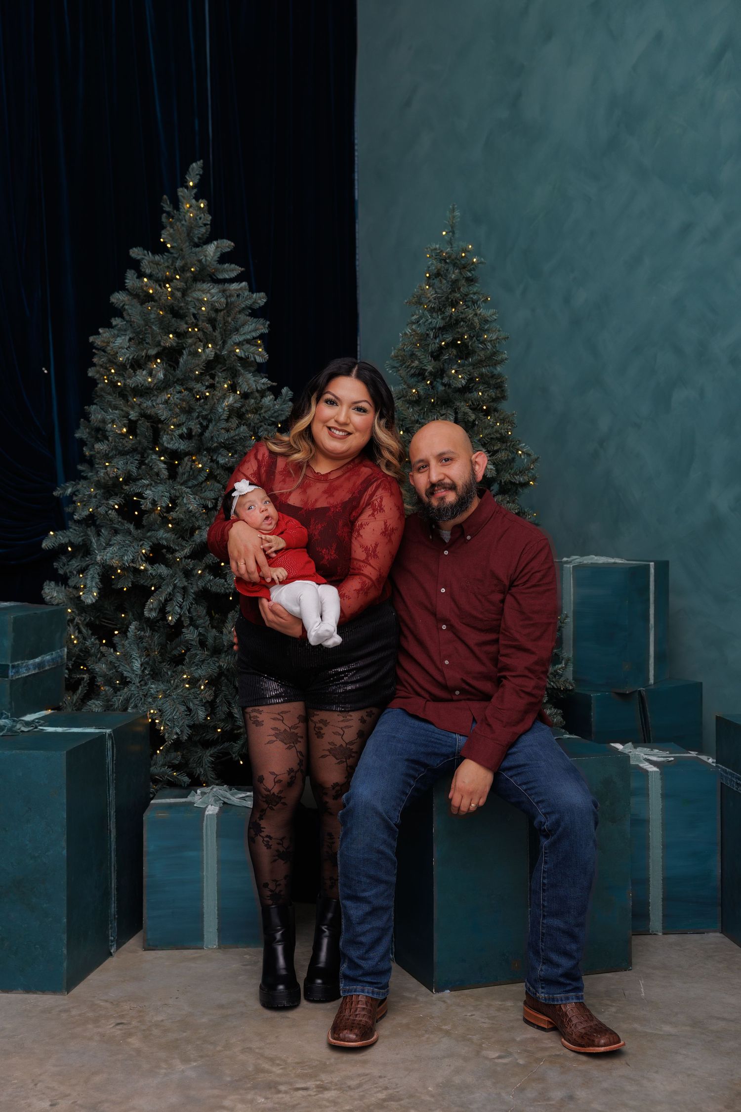 A couple sits together in a holiday photo studio setting with decorated Christmas trees and wrapped presents.