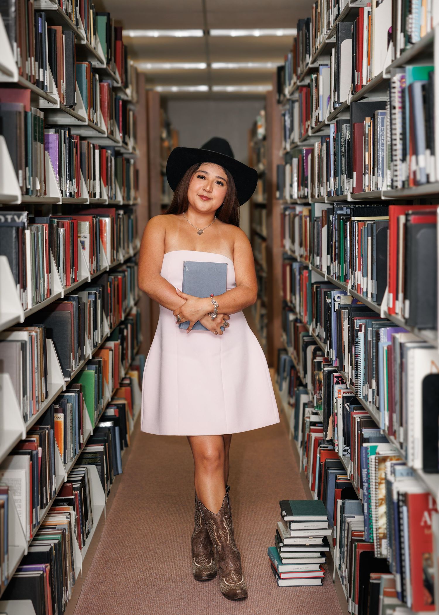Woman in white dress, black hat, and cowboy boots holding a book in a library aisle lined with bookshelves.