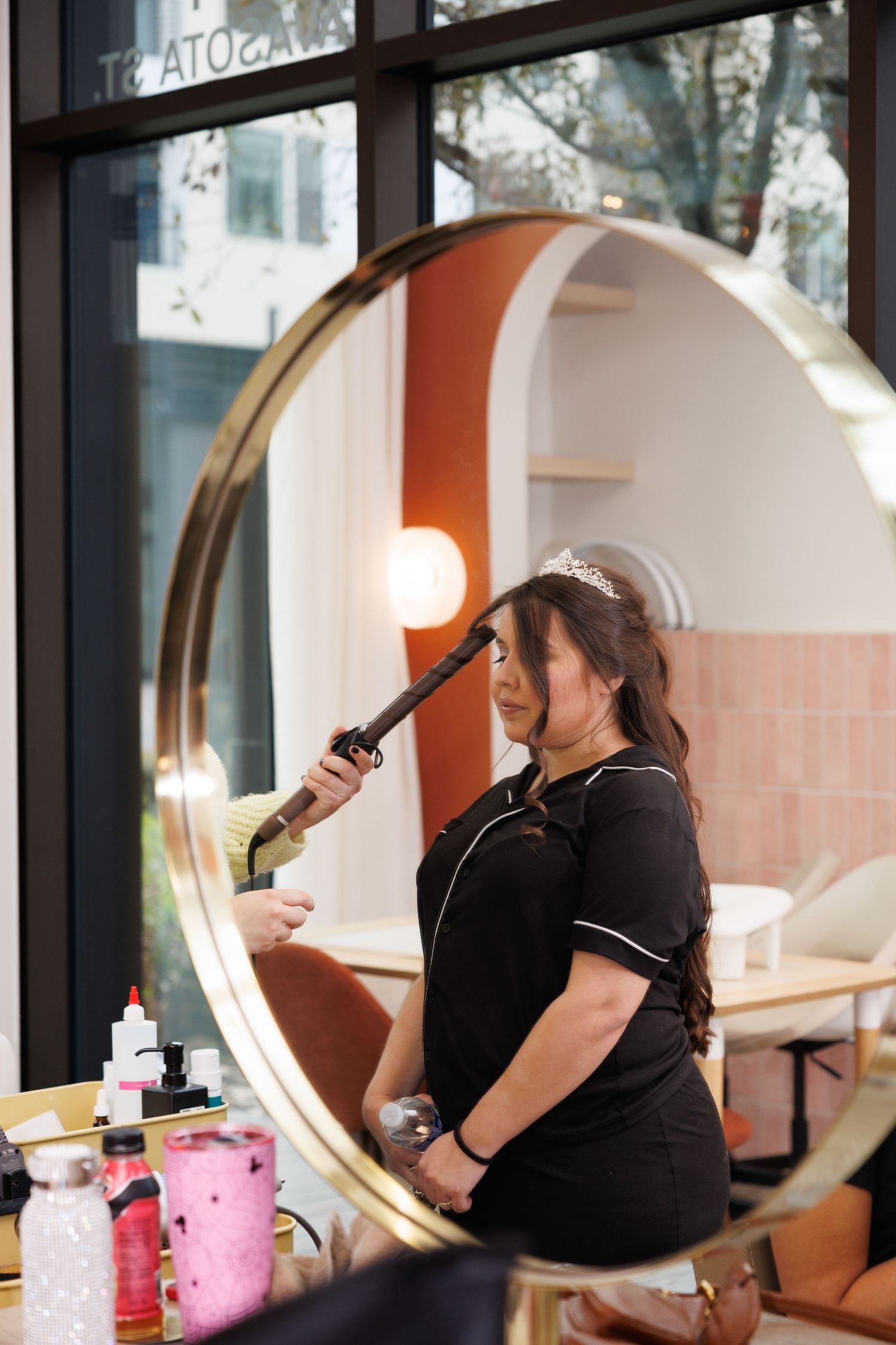 A hairstylist in a black uniform works at a salon station with a large circular mirror and beauty products on the counter.