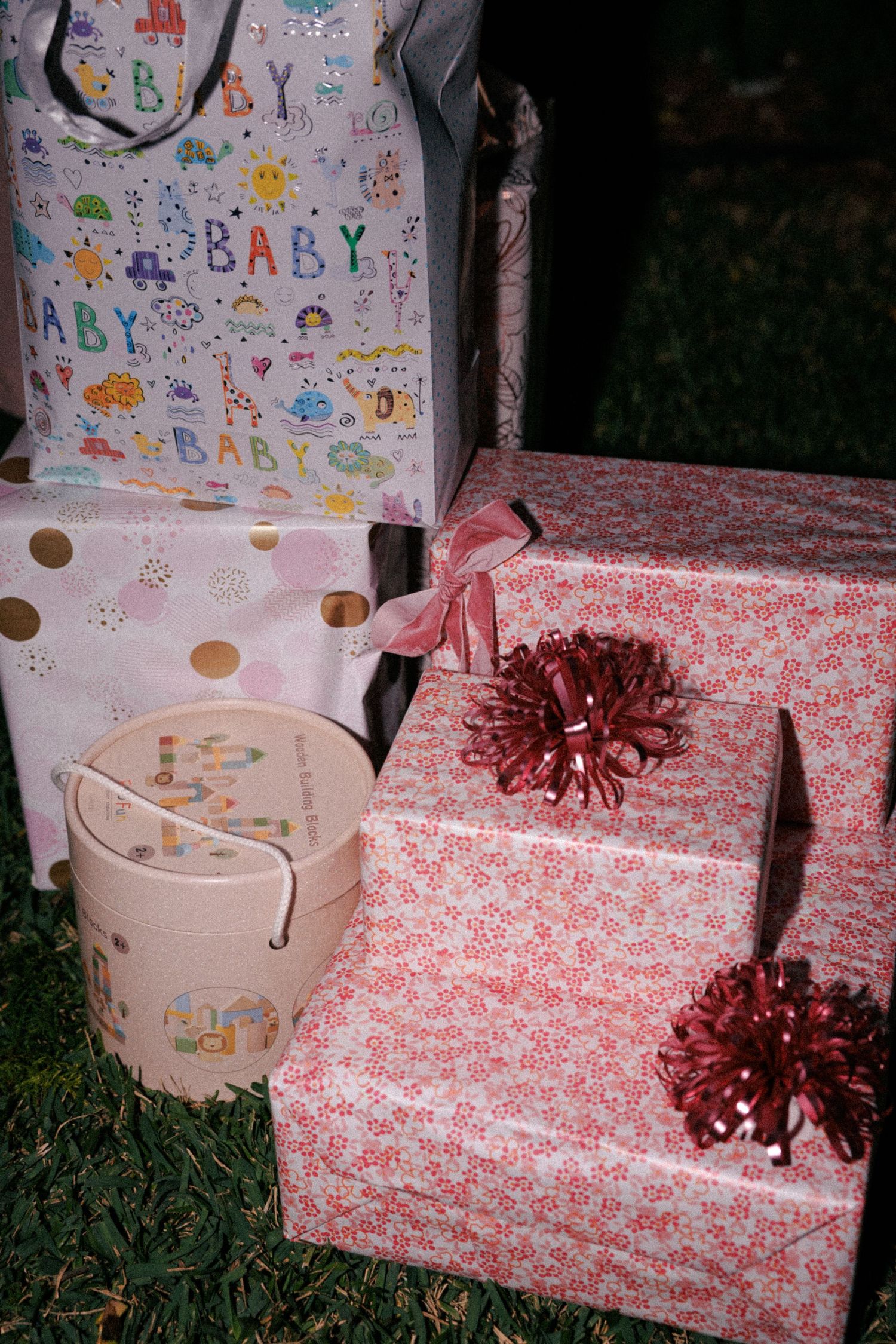 Baby shower gifts wrapped in pink floral paper with red bows and baby-themed gift bags arranged on green grass.