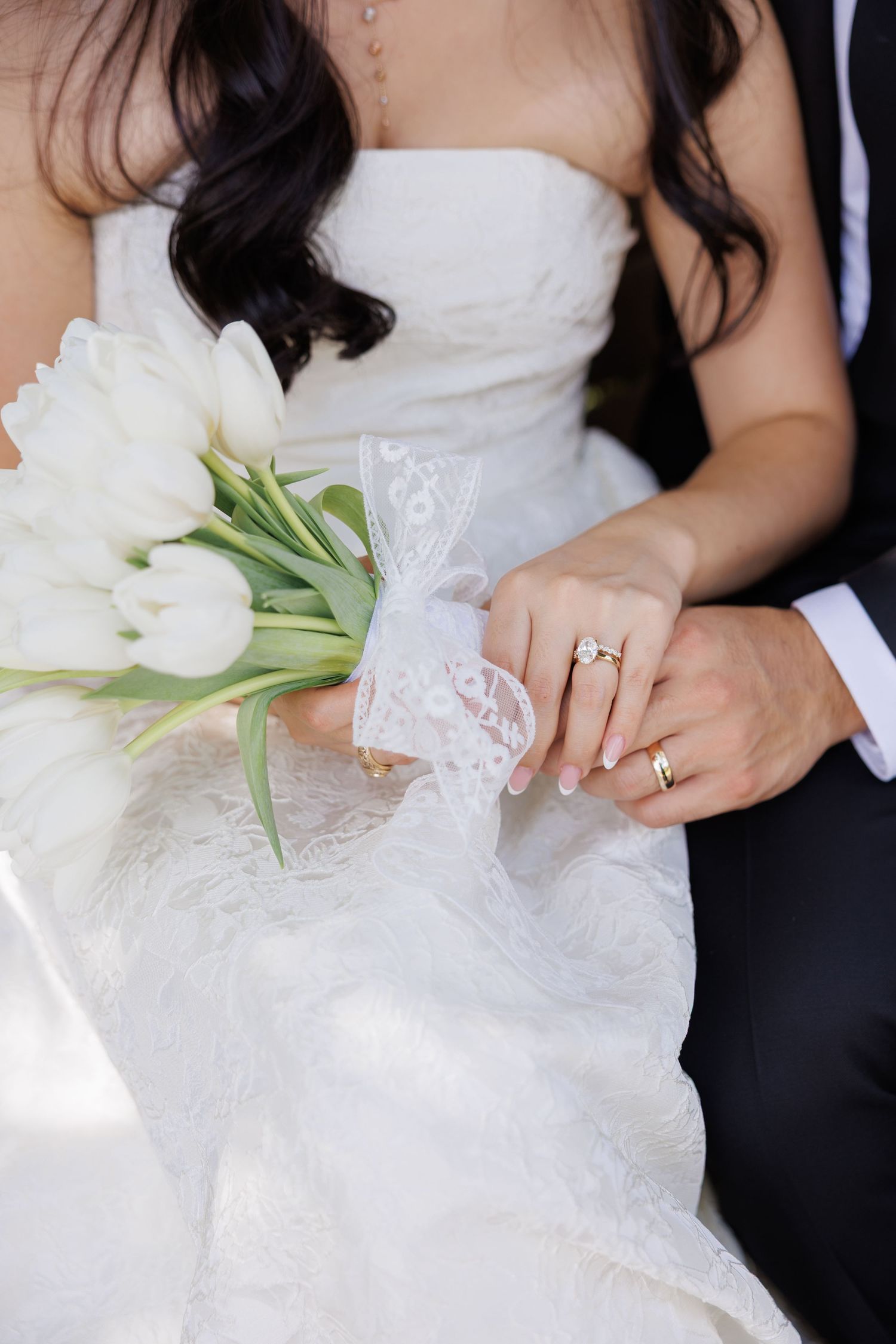 Bride in a white lace gown holds a bouquet of white tulips tied with lace ribbon as the groom's hand rests on hers, showing wedding rings.
