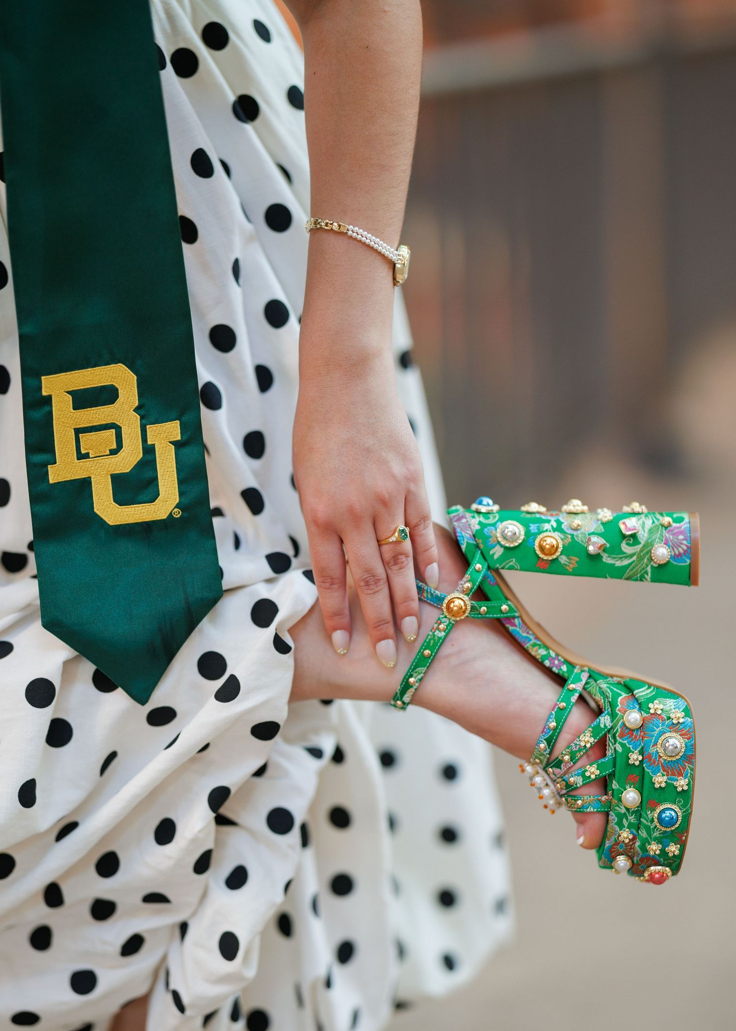Graduate in a polka dot dress and green Baylor BU stole adjusts a green jeweled platform sandal with colorful embellishments.