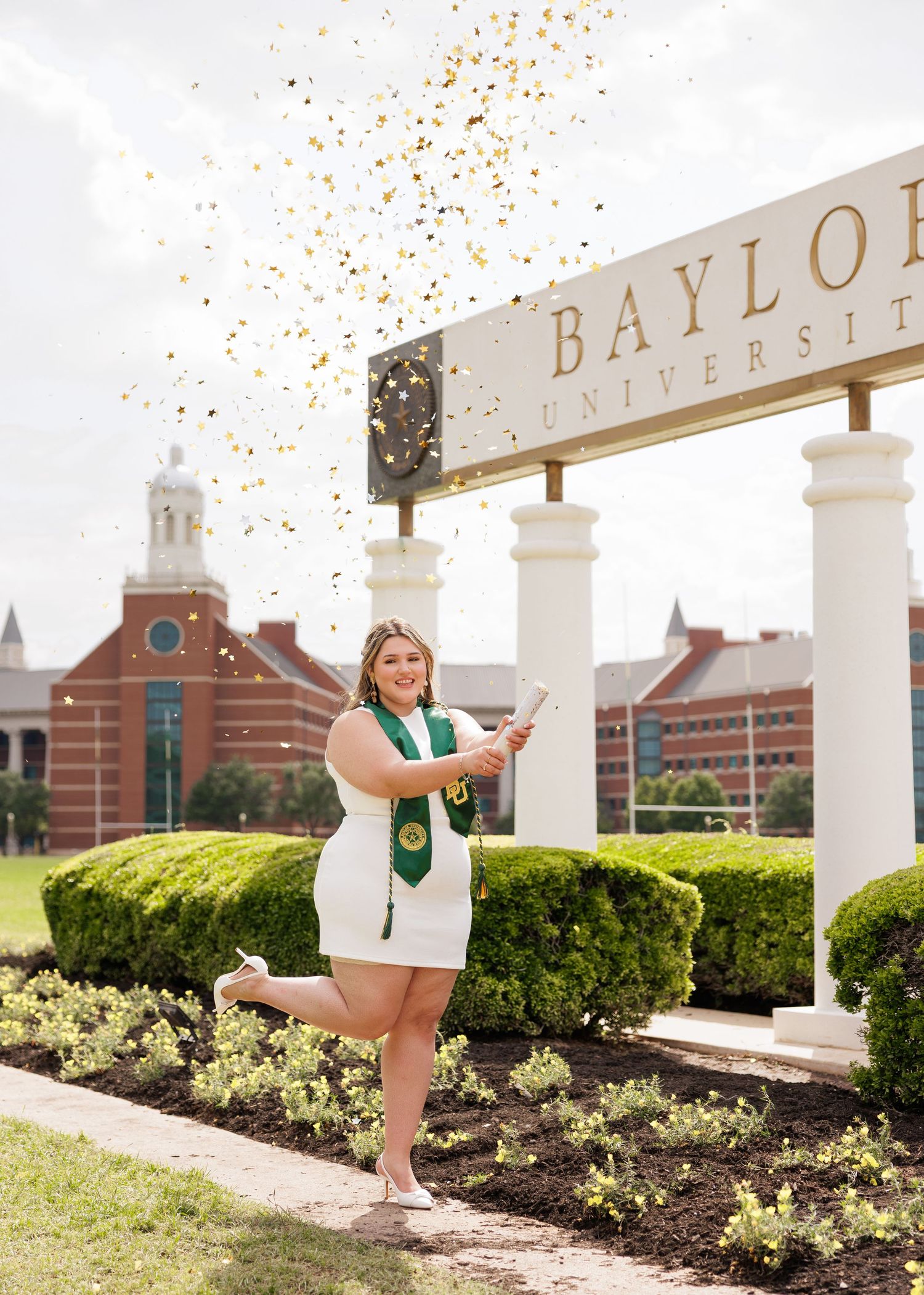 Graduate in green graduation regalia poses at different angles near Baylor University entrance columns and landscaping.