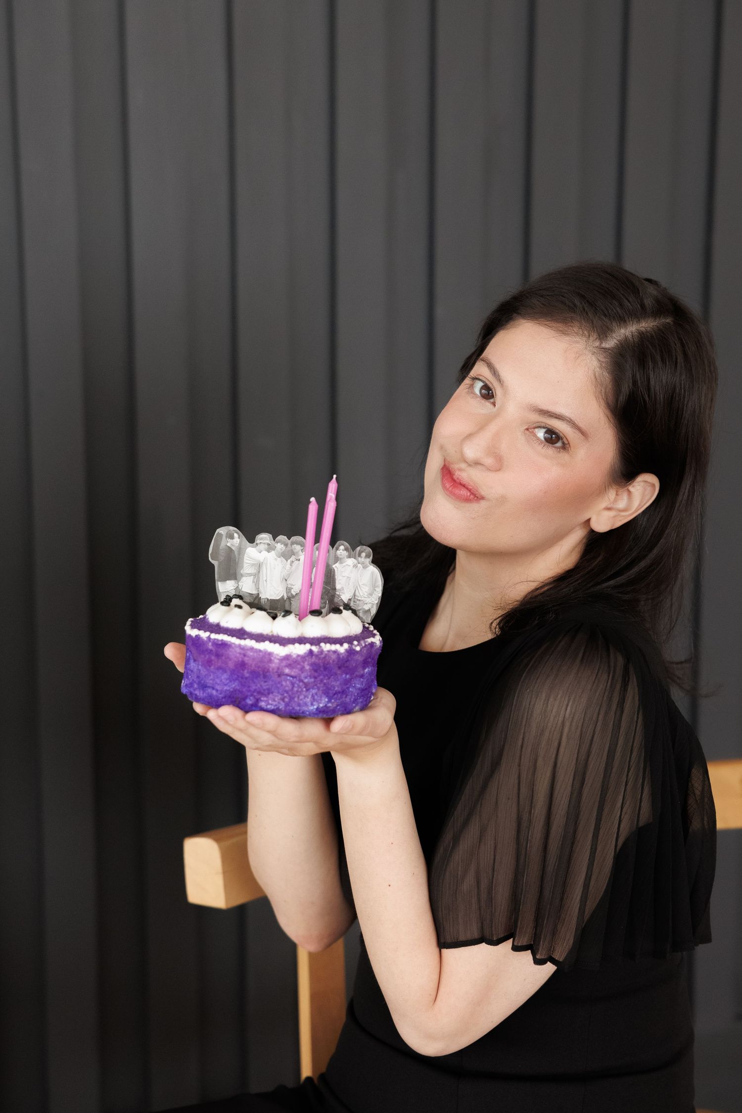 Someone in a black shirt holds up a small purple birthday cake with candles and white frosting decorations against a dark background.