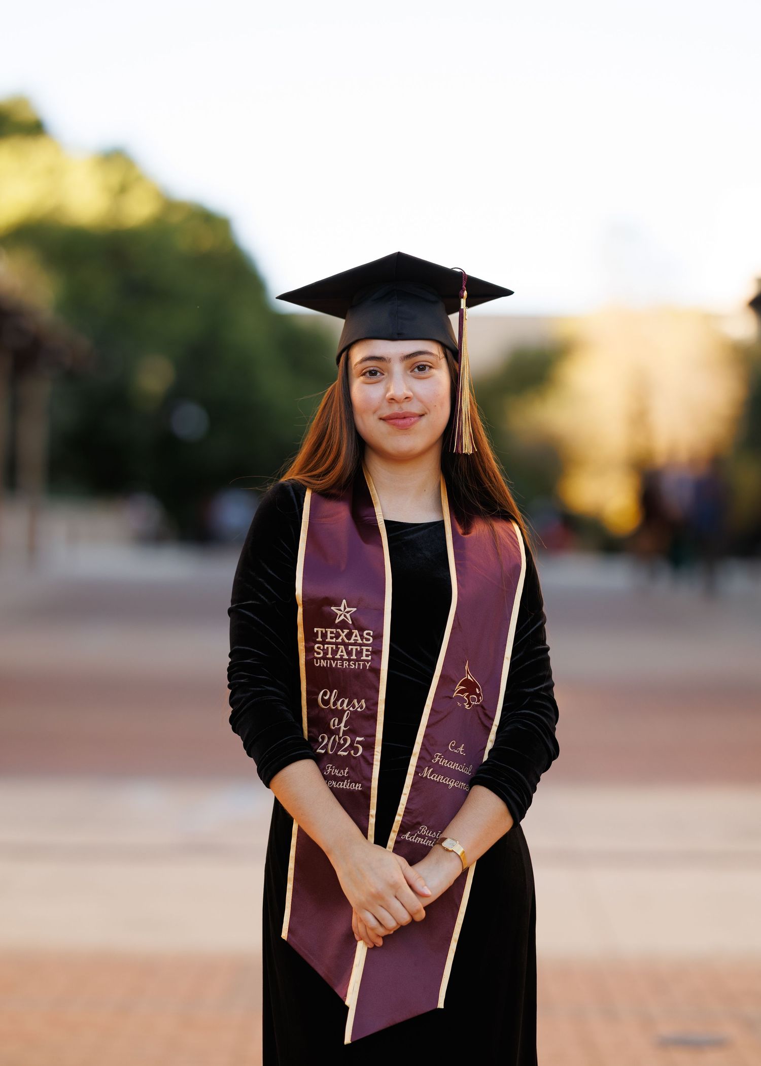 Graduate in black cap and gown holds diploma and stole during outdoor commencement ceremony at sunset.