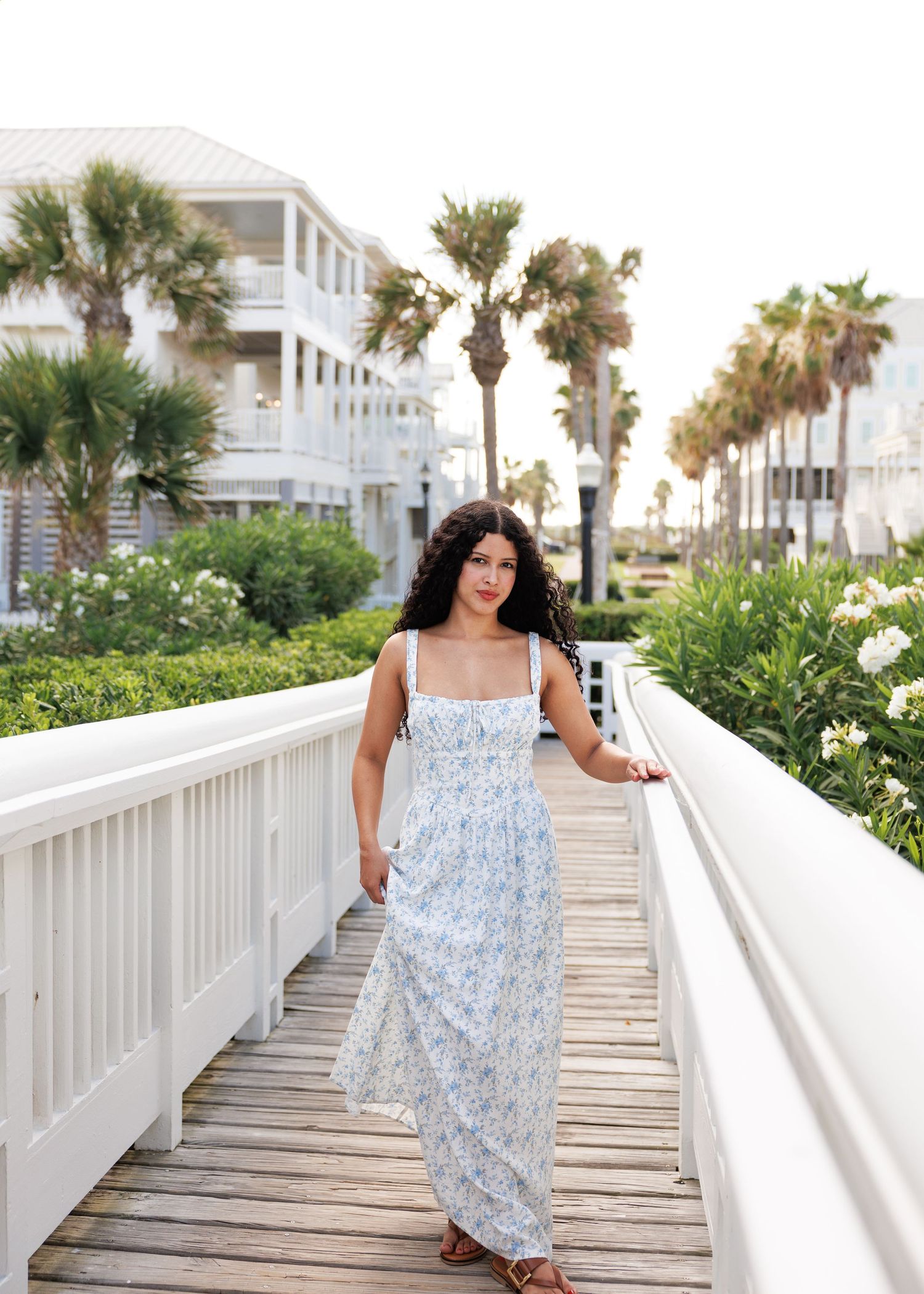 A person in a flowy white and blue floral dress walks along a beachfront boardwalk lined with palm trees and coastal homes.