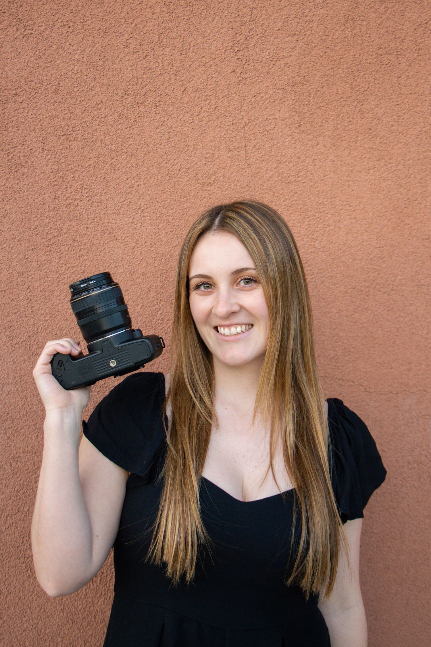 A photographer in a black top holds a professional camera against a terracotta colored wall.