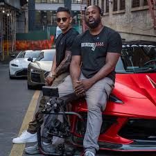 Two people pose with luxury sports cars and a motorcycle on an urban street.