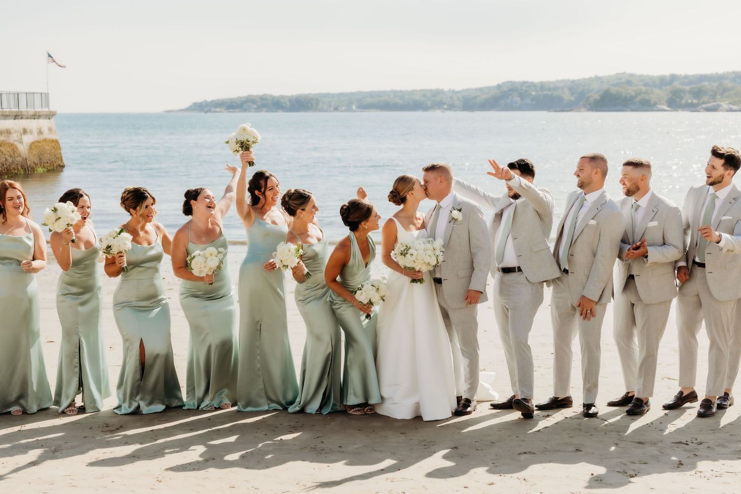 Wedding party in sage green dresses and light colored suits standing in line against ocean backdrop.