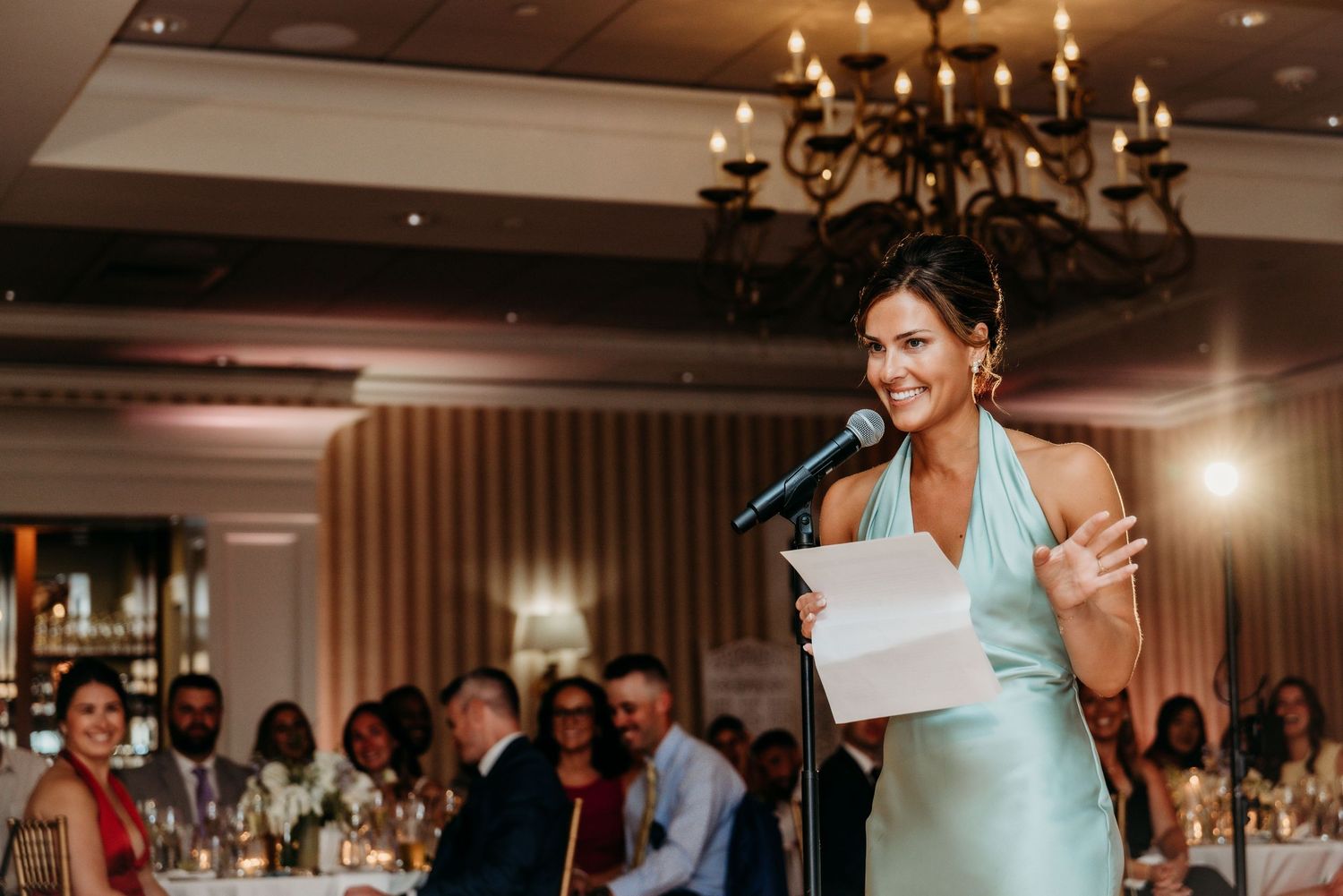 A person in a turquoise dress delivers a speech at an elegant wedding reception with a chandelier overhead.